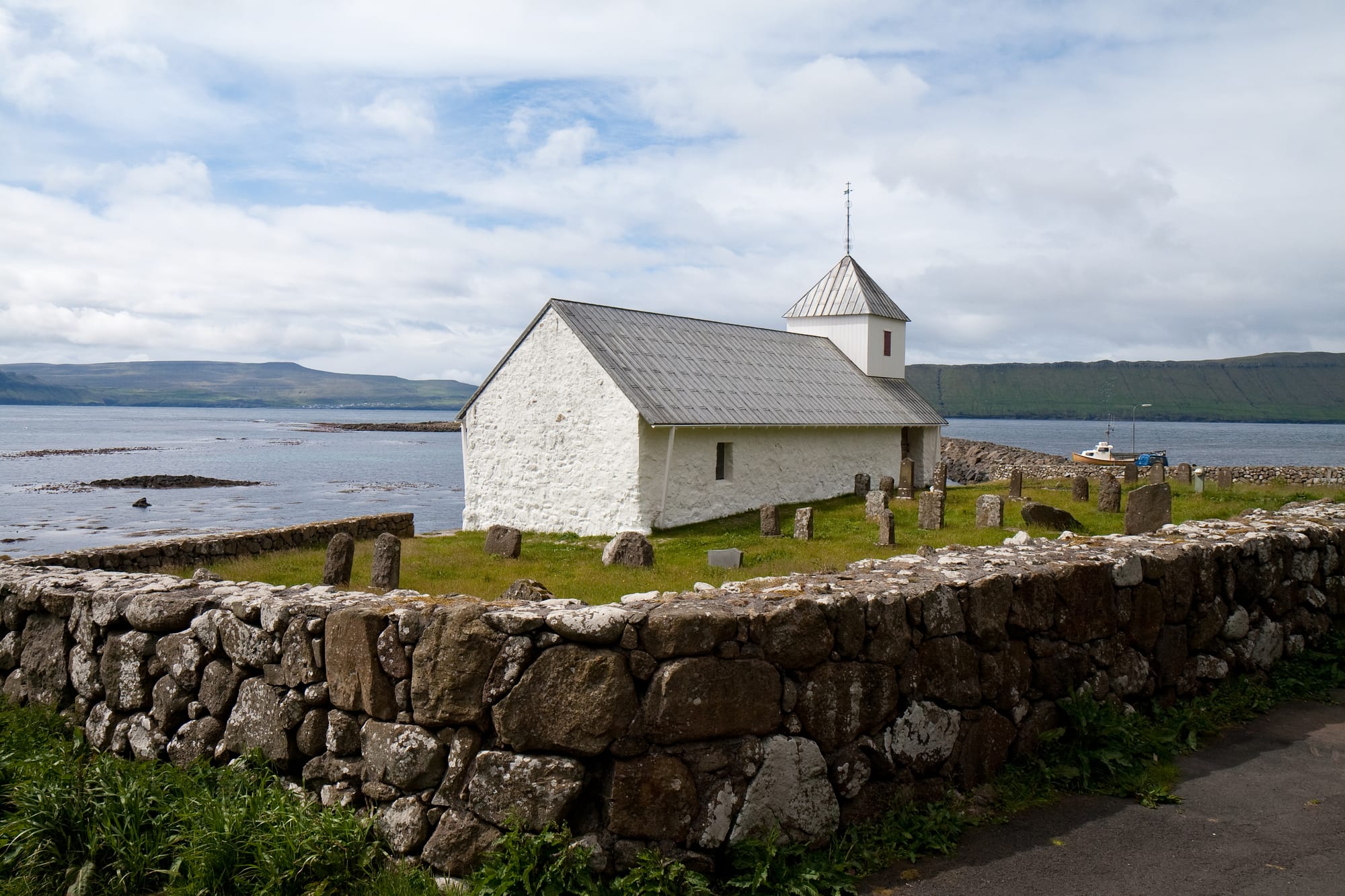 A small whitewashed stone church with a grey roof and a little white bell tower, standing in a churchyard with old headstones behind a mossy dry-stone wall, beside the sea