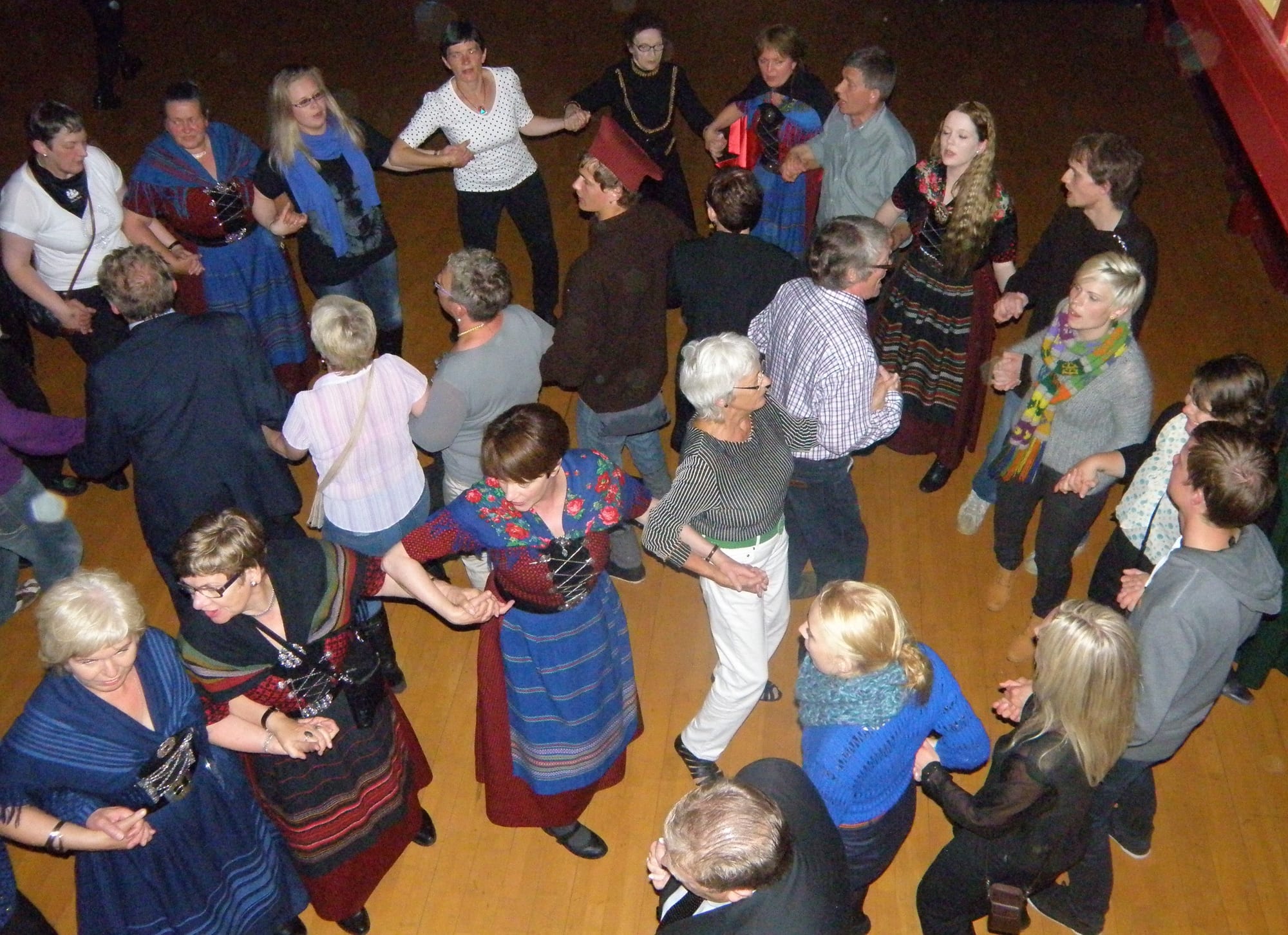 Seen from above, a crowd of men and women, several in red, blue and black Faroese national dress, hold hands in a winding line as they dance on a wooden floor