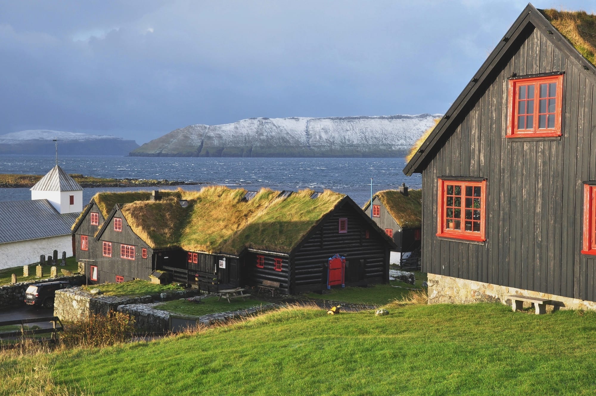 Turf-roofed black timber farm buildings beside a stone church on a green shore