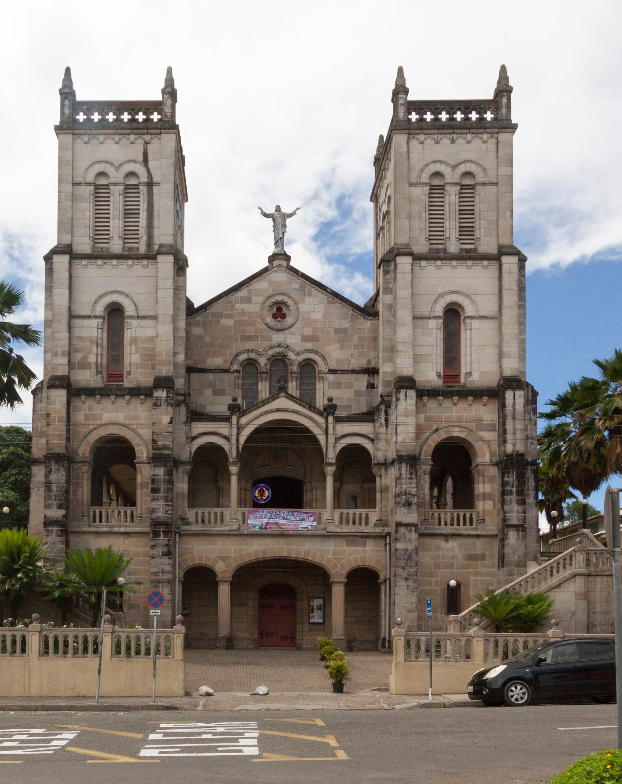 The stone front of a cathedral with two square towers, arched windows and a white statue of Christ with outstretched arms on the central gable, above an arched entrance porch and a low wall