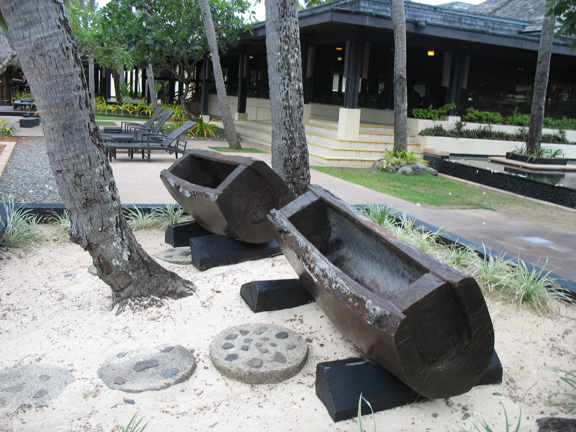 Two large hollowed-out wooden slit drums resting on black supports on white sand between palm trunks, with a low building and sun loungers behind