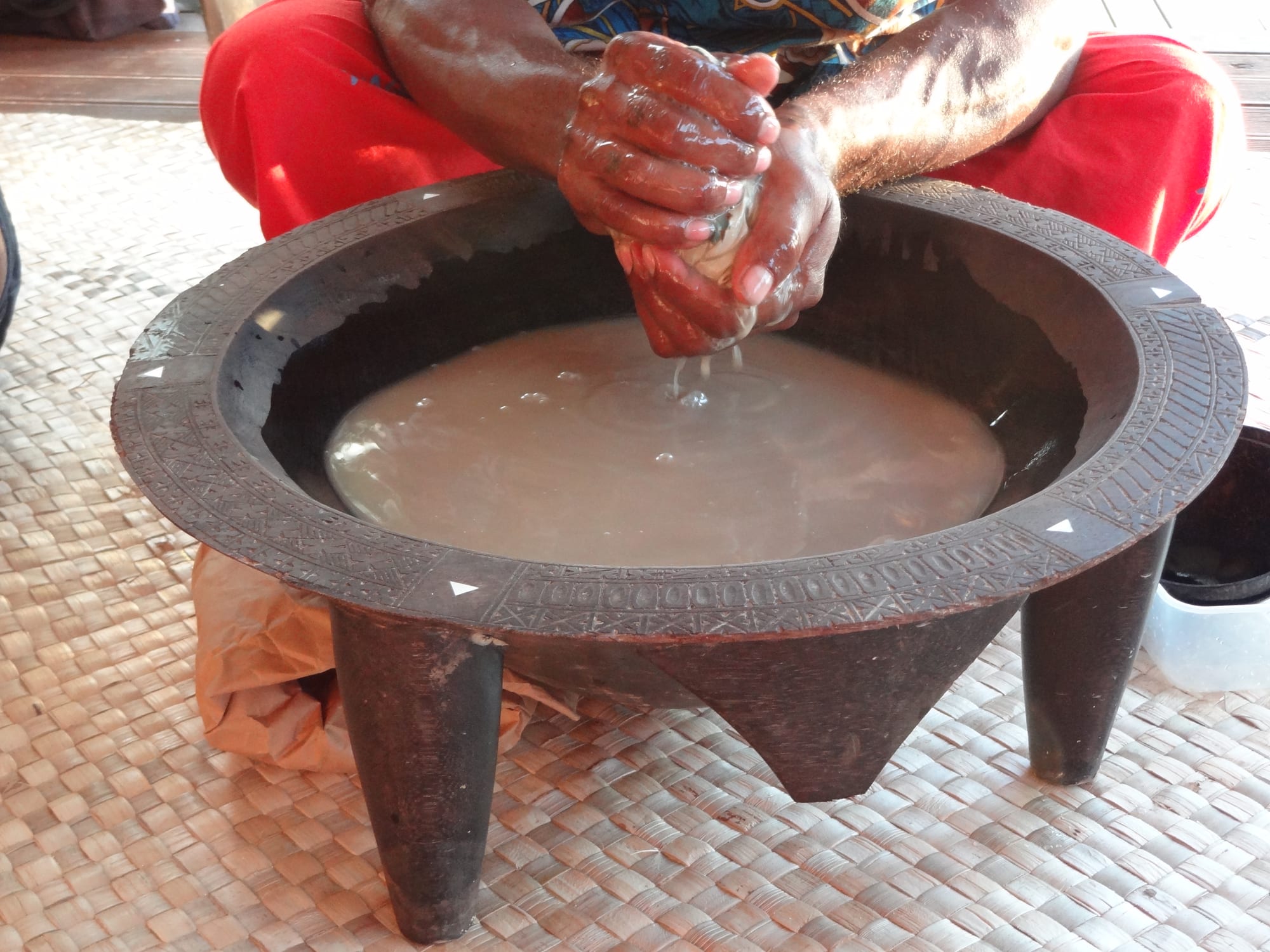Two hands squeezing a cloth bundle of kava into a large carved wooden bowl on short legs, filled with cloudy brown liquid, set on a woven mat