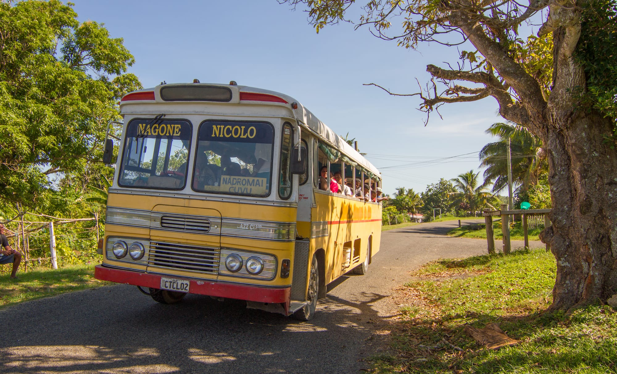 An open-sided yellow and white Fijian bus carrying passengers along a rural road near Cuvu Village, Viti Levu