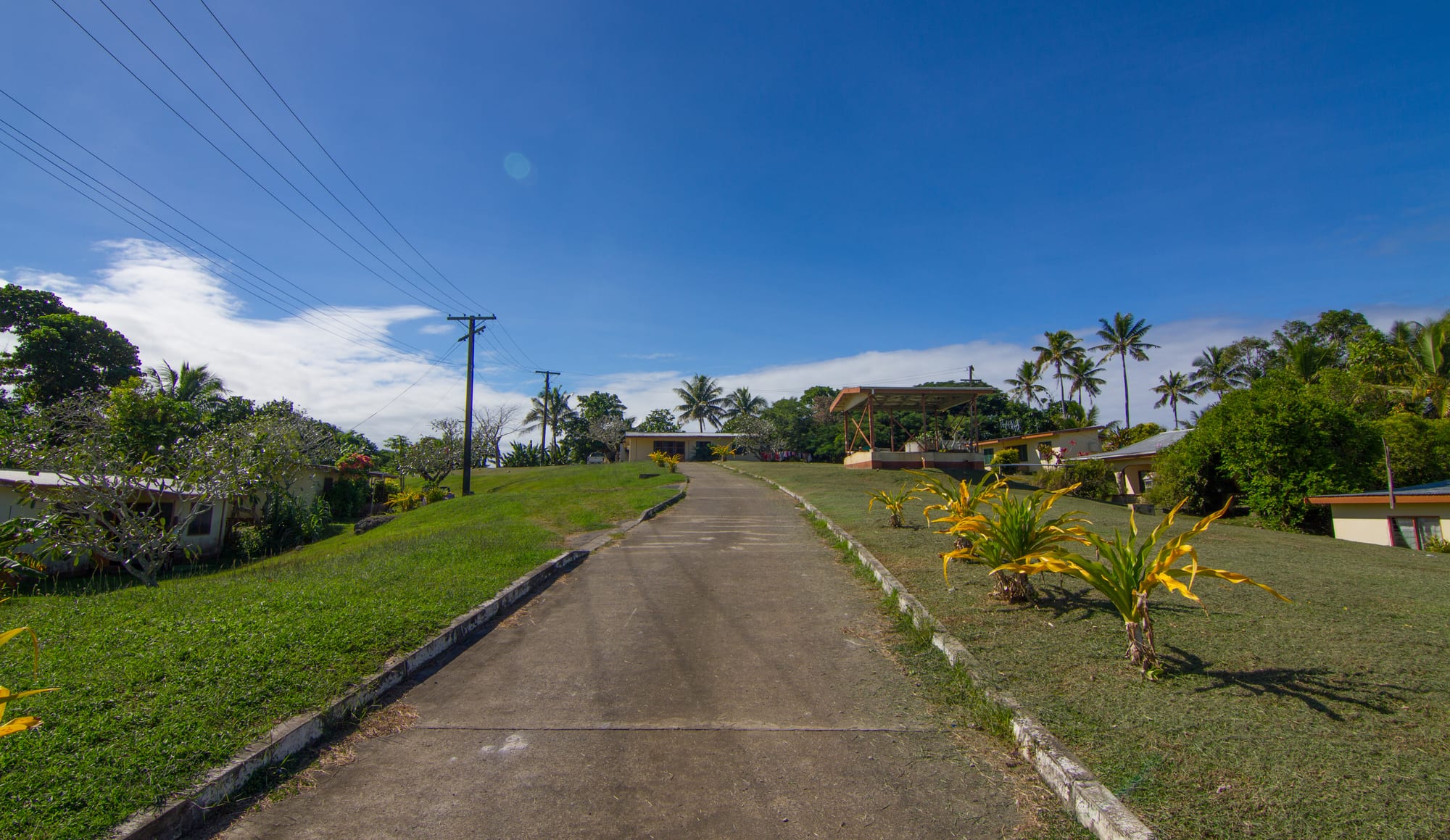 A paved path leading up a grassy hillside to houses and an open-sided community hall in an iTaukei village in Fiji