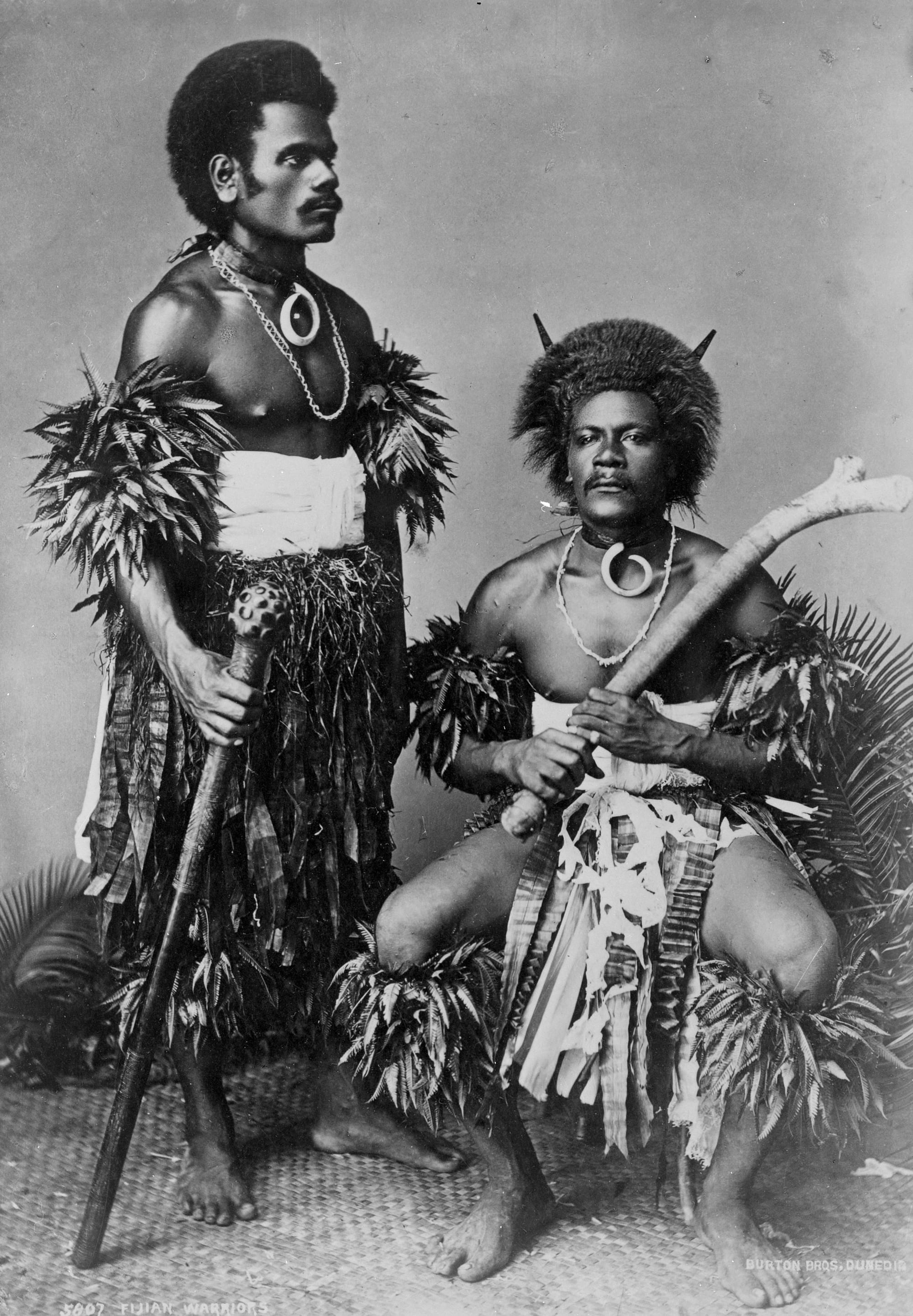 Two Fijian men in the 1880s in barkcloth, holding large war clubs, photographed in a studio