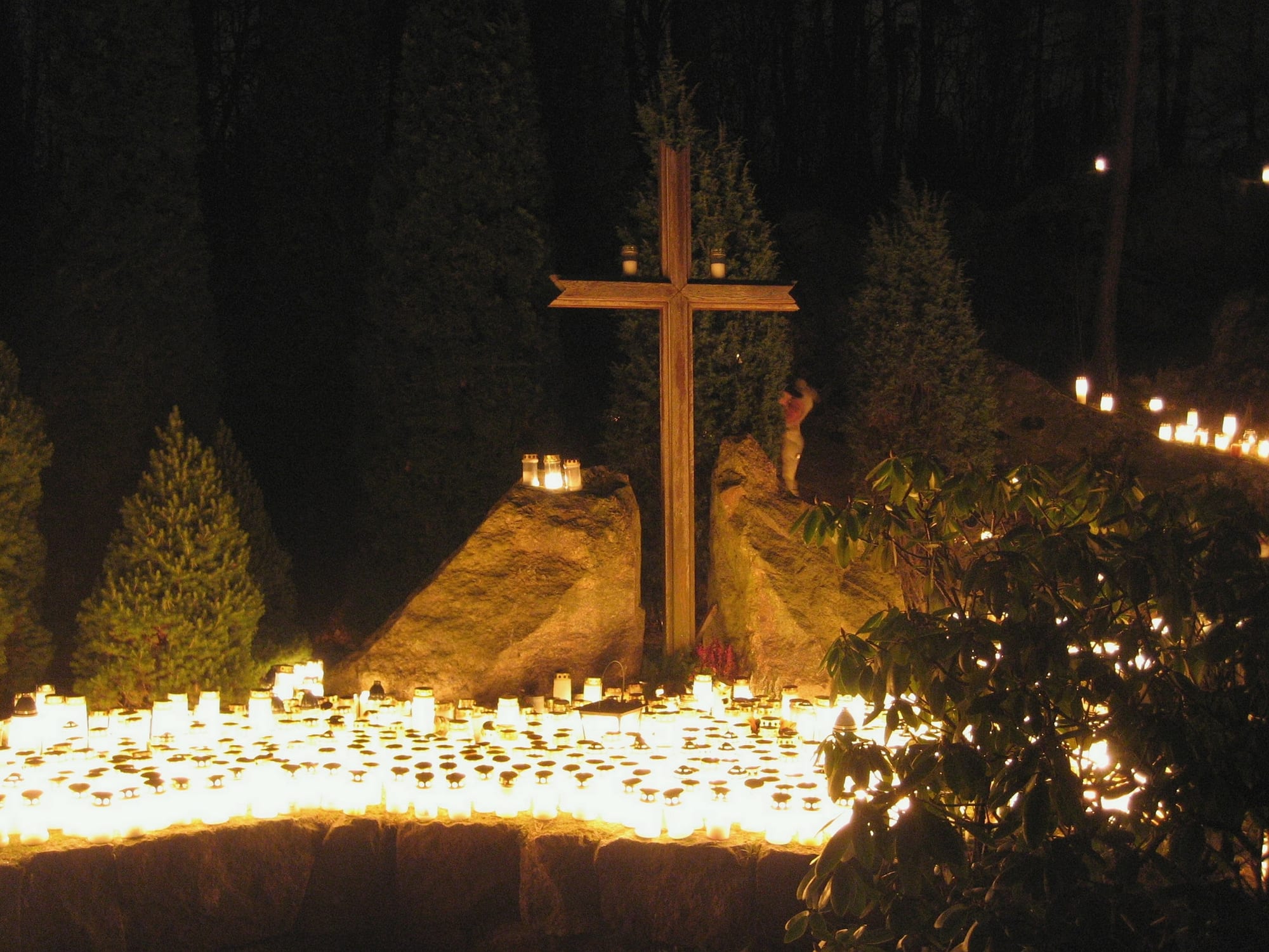 Hundreds of small white candles in glass holders glowing on the ground around the base of a tall wooden cross between two rocks, with dark fir trees behind
