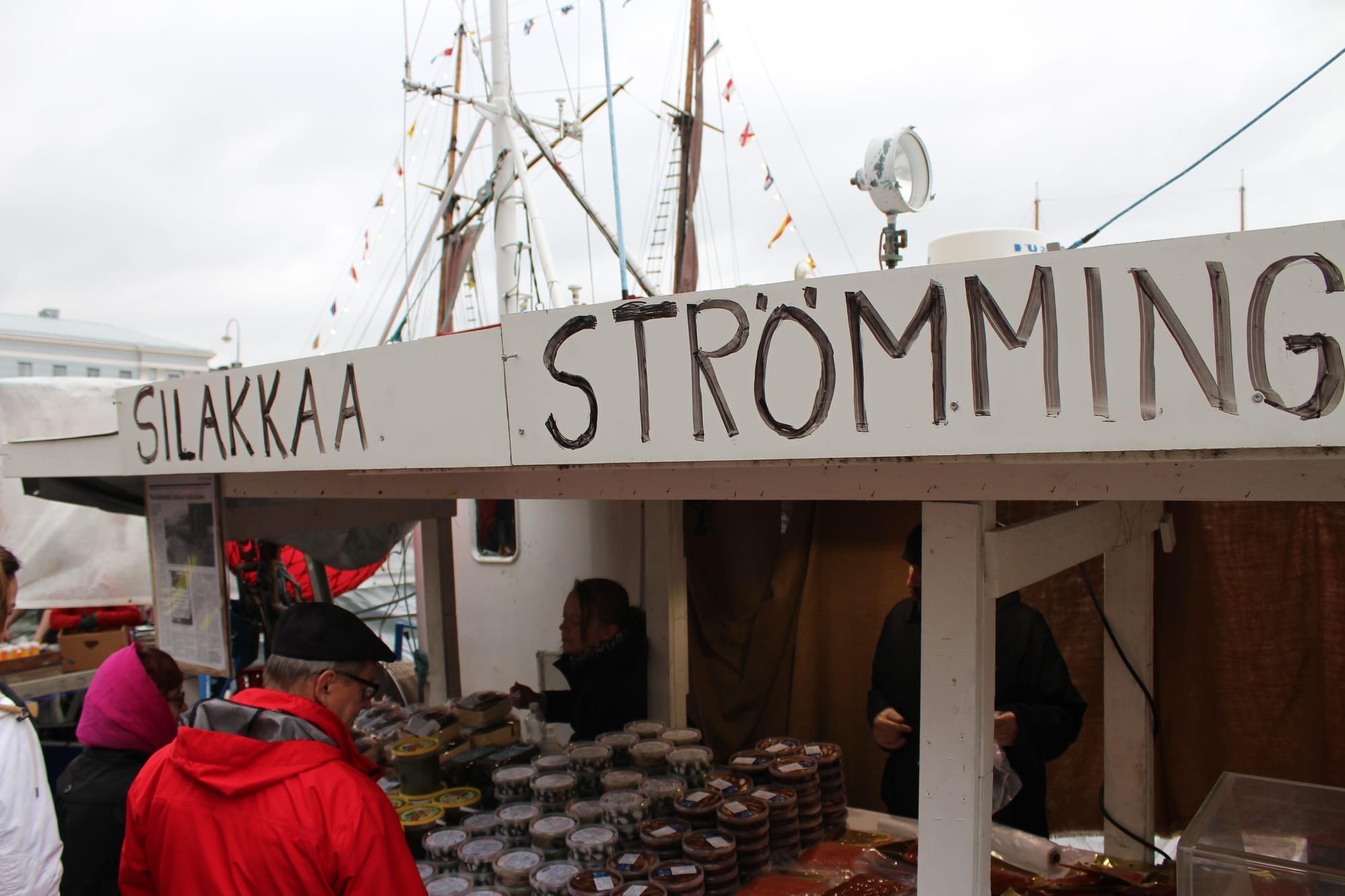 A market stall at Helsinki's harbourside Baltic herring market, selling tubs of pickled herring, with sailing ships moored behind