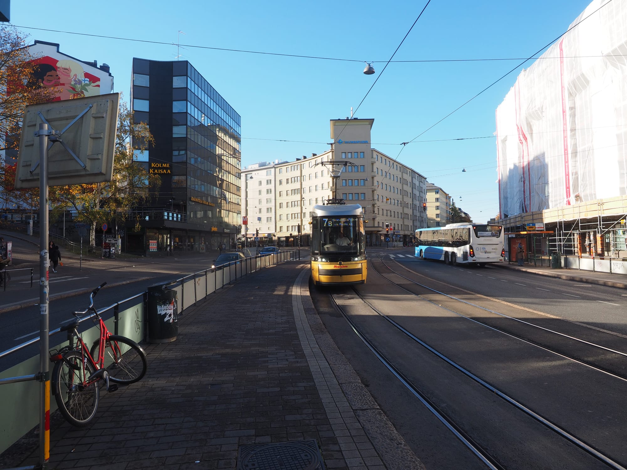 A yellow Helsinki tram rounding a curve on a city street, with a bus, cyclists, and apartment buildings behind