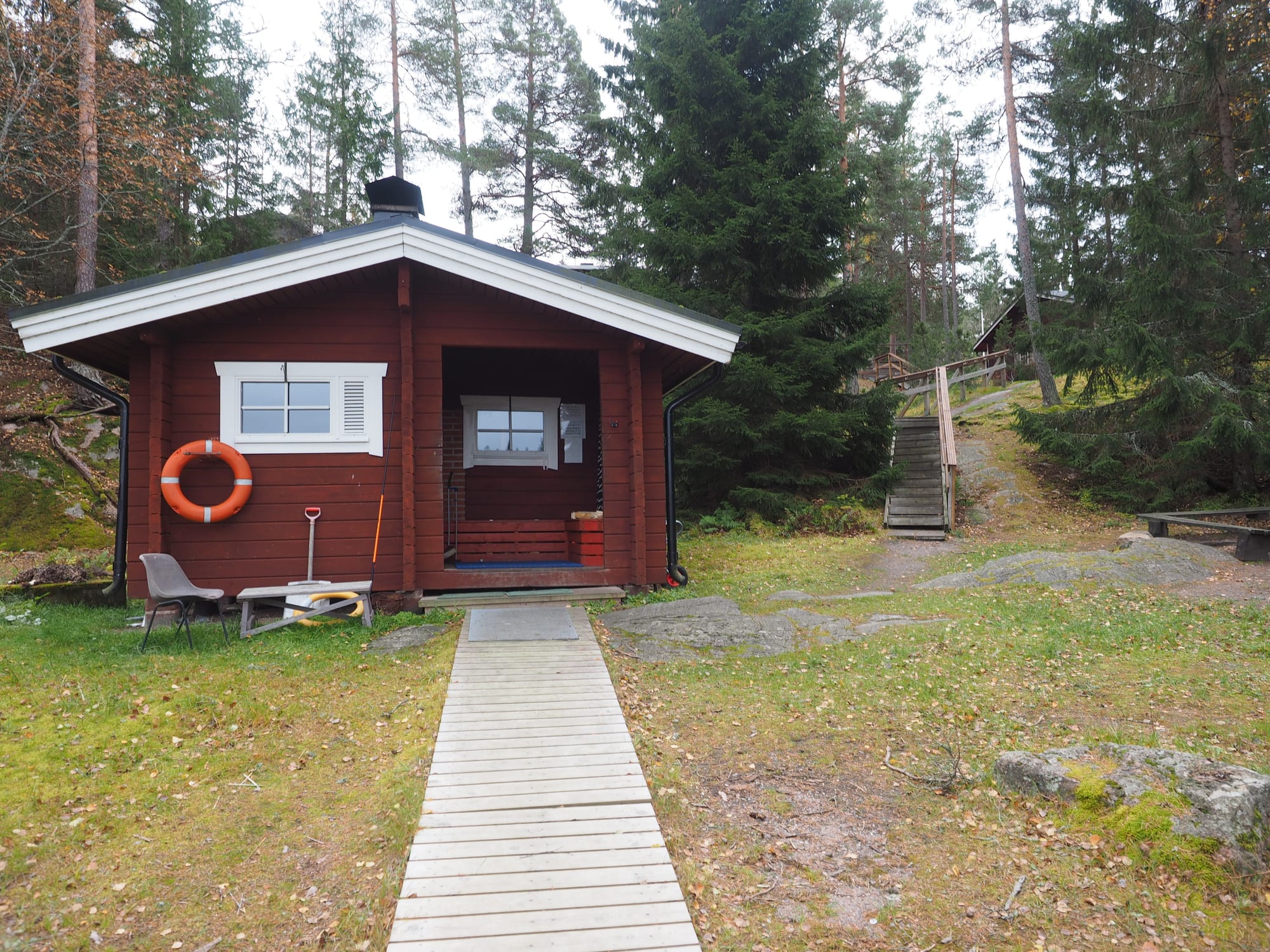 A small red wooden sauna building with a wooden walkway leading to it on a rocky shore in Kirkkonummi, Finland