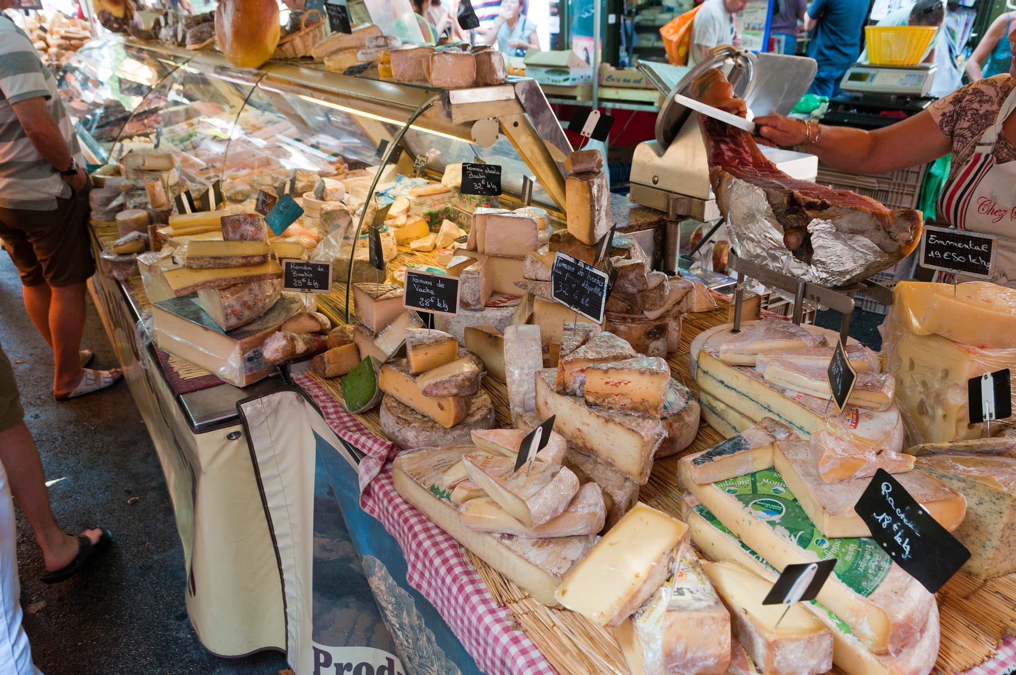 A French market cheese stall piled high with wedges and wheels of different cheeses, price tags on small blackboards, shoppers behind