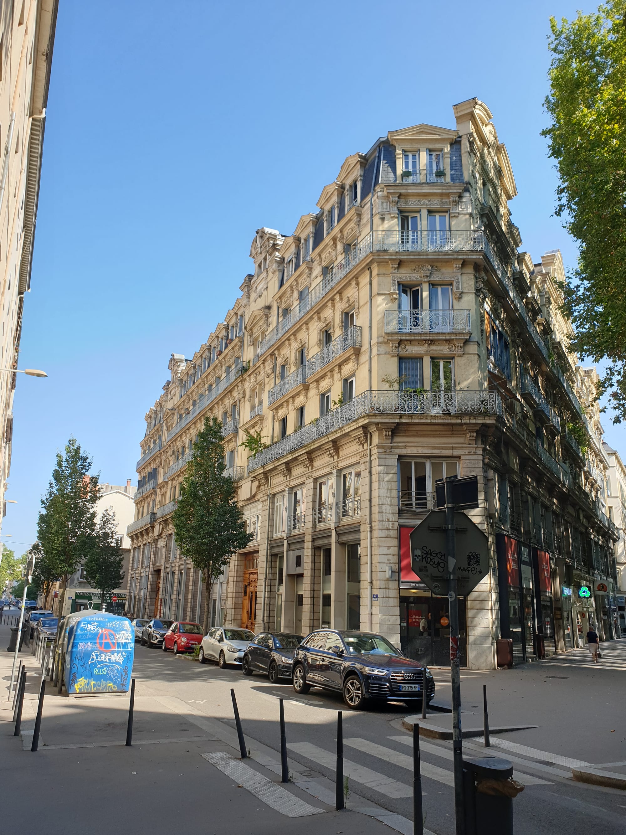 A tall cream-coloured Haussmannian apartment building with wrought-iron balconies on a street corner in Lyon