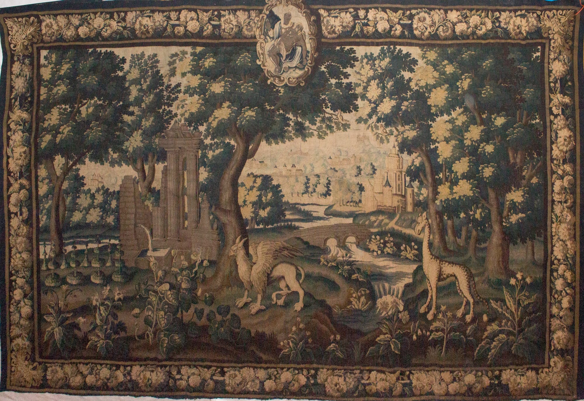 A large woven tapestry in greens and browns showing a wooded landscape with a giraffe and a griffin in the foreground, ruined columns, a river and a castle, inside a border of flowers