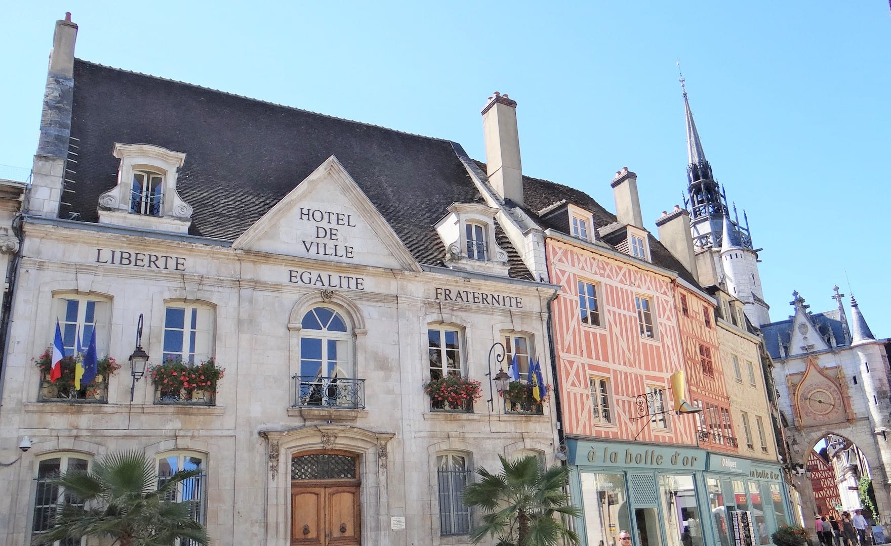 The pale stone front of a town hall with the words Hotel de Ville on its central gable and Liberte, Egalite and Fraternite carved across the facade, with flags and flower boxes at the windows