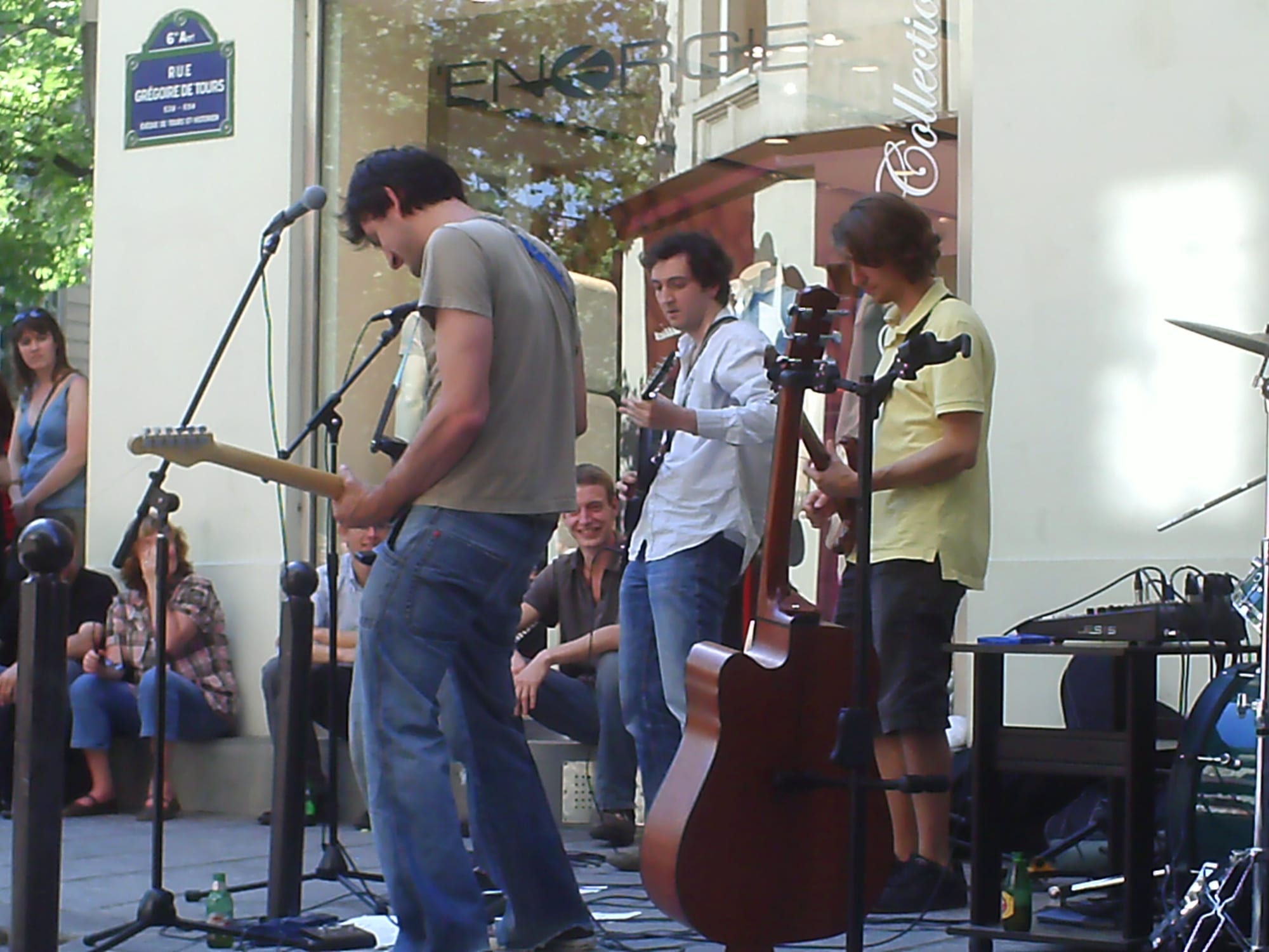 A small band with electric and acoustic guitars plays on a pavement outside a shop while people sit on a ledge behind them listening