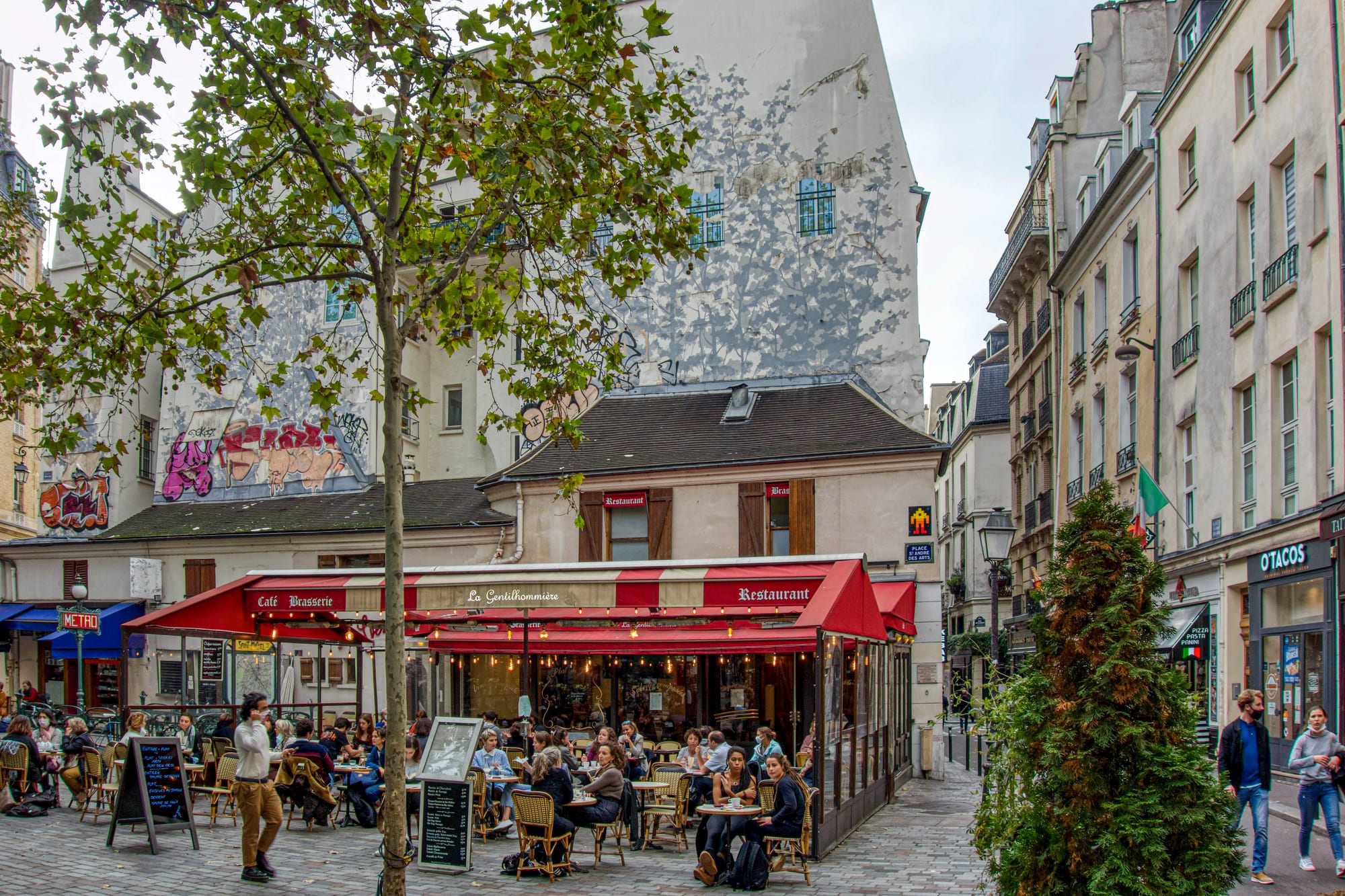 A café-brasserie with a red and white striped awning and people seated at small round tables on its terrace, with tall pale buildings and a tree around a paved square