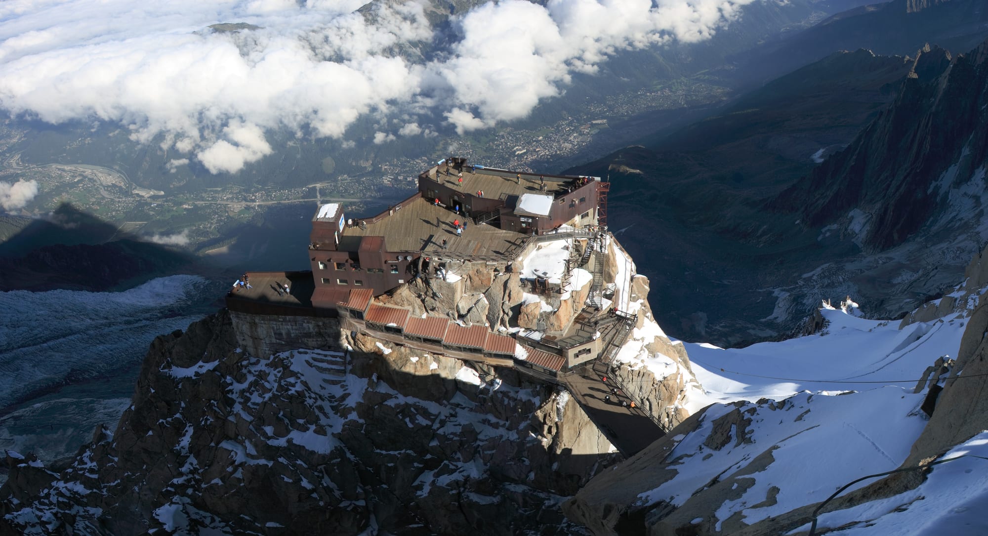 A rocky mountain spire topped by a cable-car station, with snow peaks all around