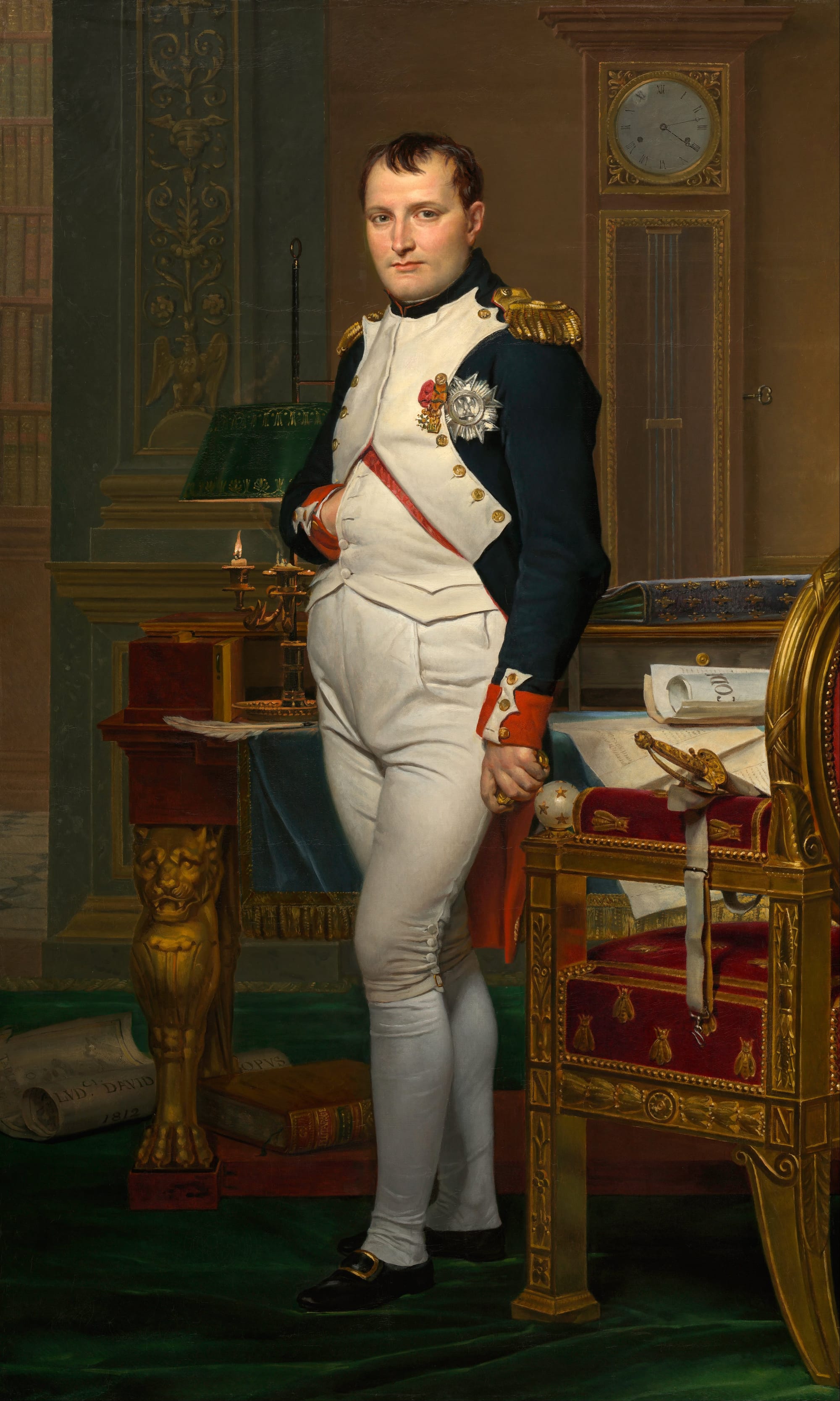 A full-length portrait of Napoleon in a military uniform standing by a desk in his study