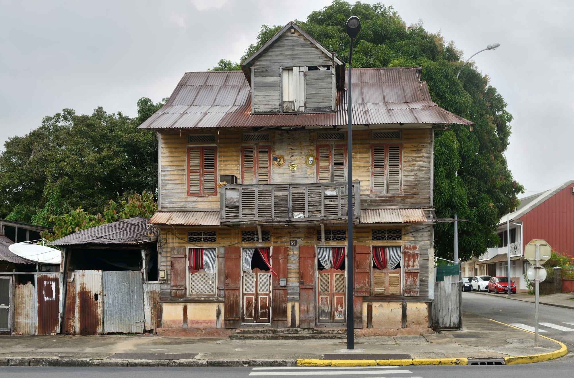 A weathered two-storey wooden Creole house with a corrugated roof and shuttered windows on a street corner in Cayenne