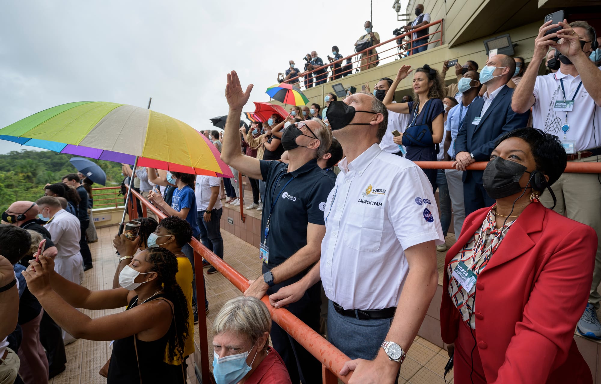 A crowd of people, some holding colourful umbrellas against rain, watching a rocket launch from a viewing gallery at the Guiana Space Centre