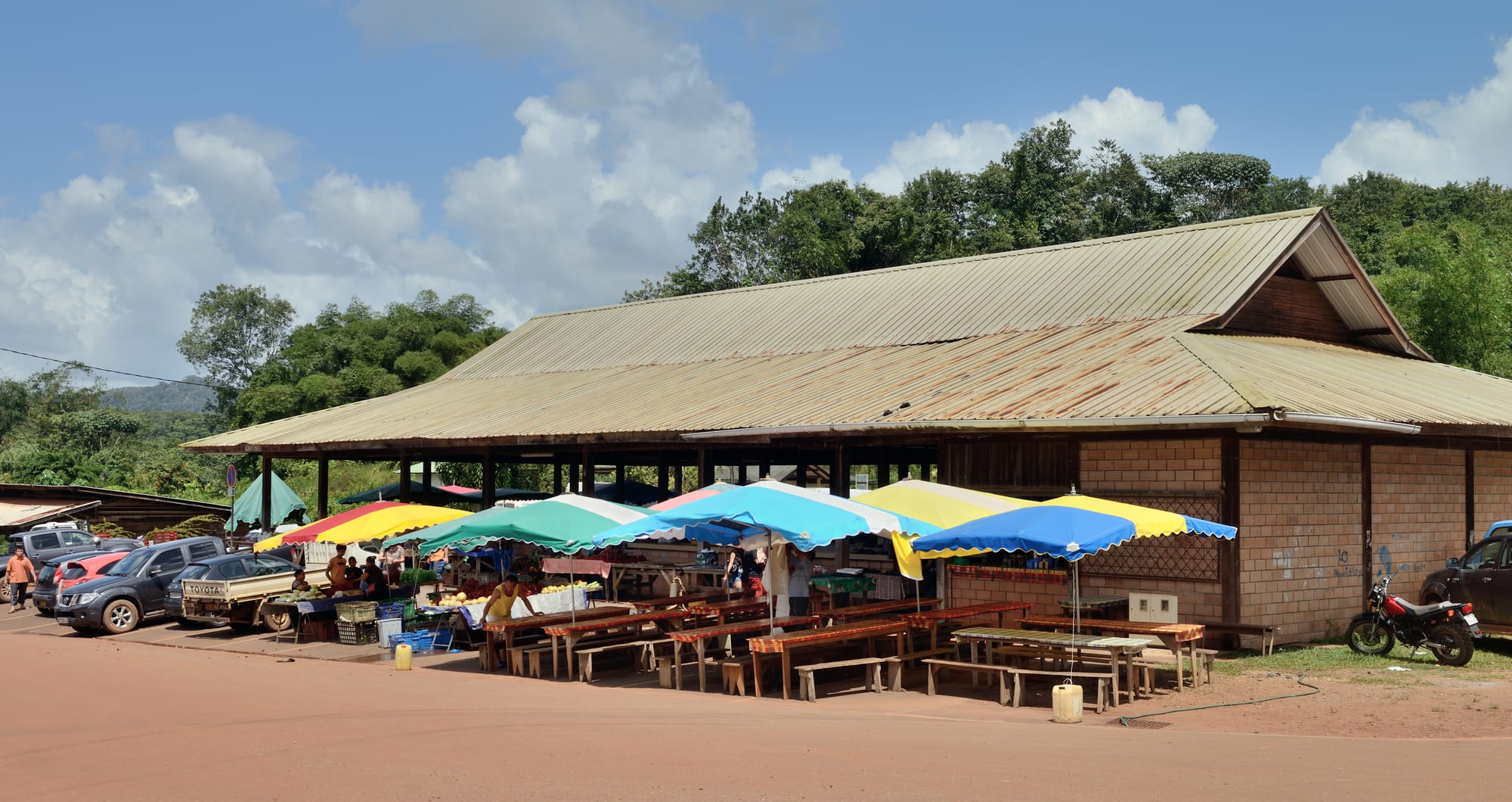 A covered market with colourful umbrella stalls, parked cars and motorbikes at Cacao village, French Guiana, surrounded by rainforest