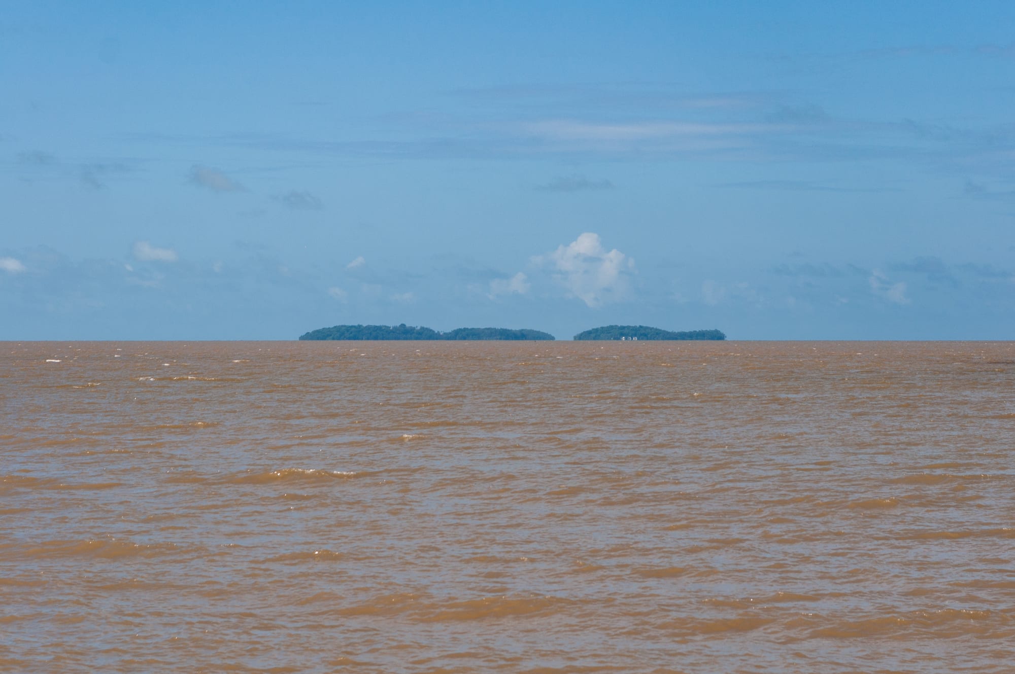 Three low forested islands in a calm blue sea seen from a rocky shore