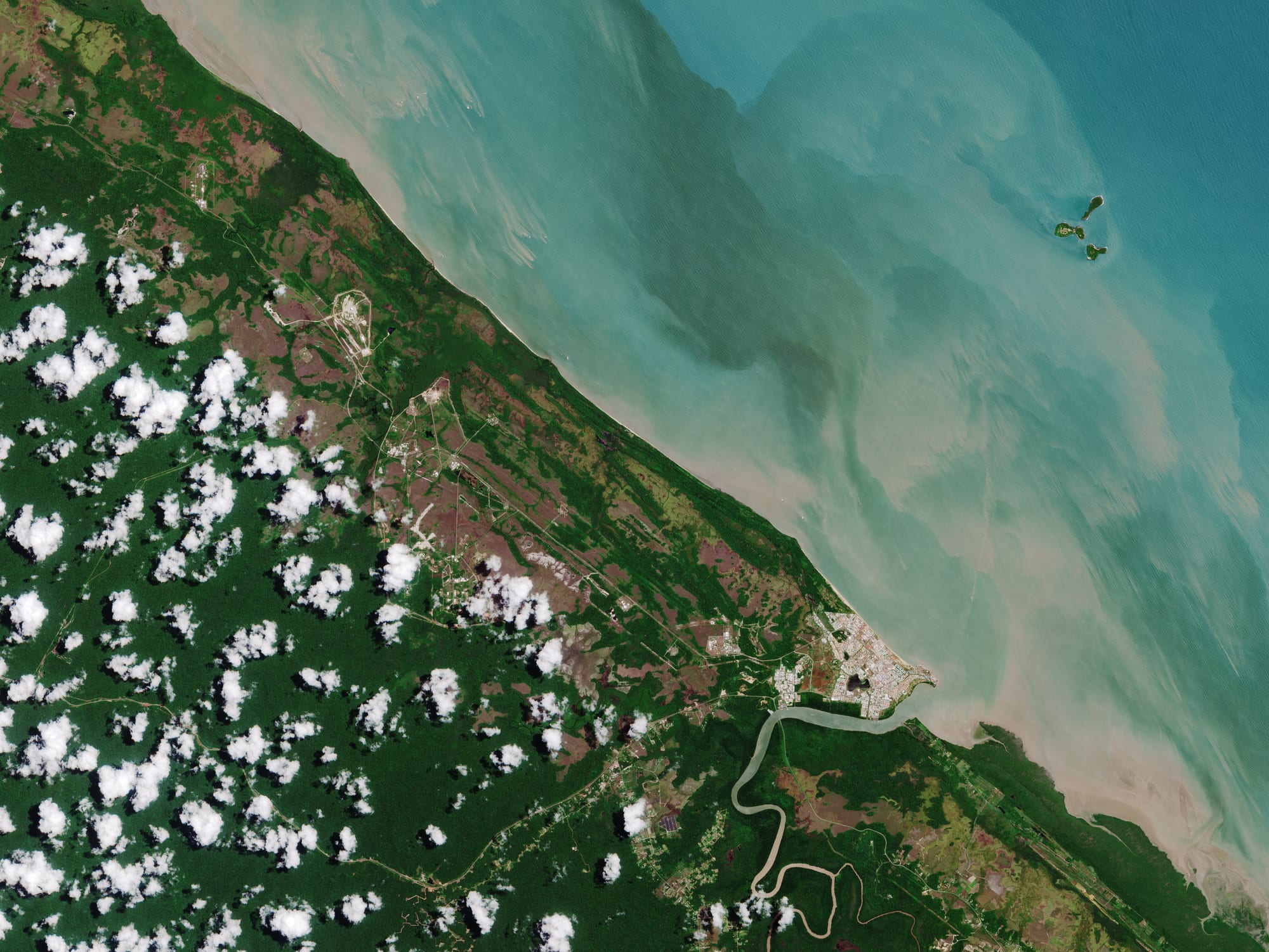 A rocket launch tower and assembly buildings in a clearing surrounded by rainforest and sea
