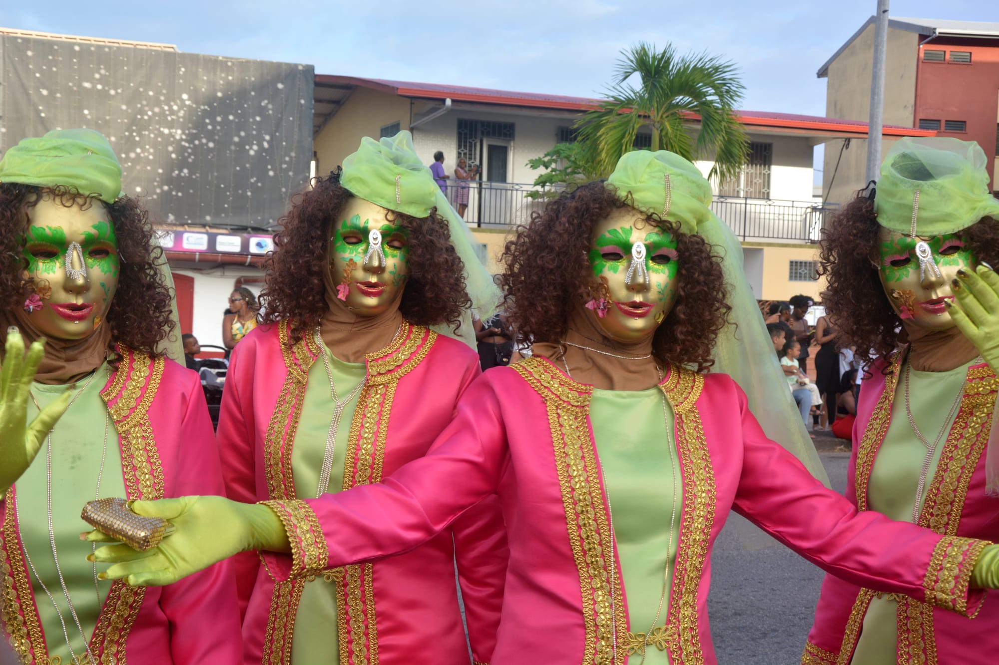 Four masked Touloulous in matching pink and pale green costumes with gold trim, gloves, painted masks and green headscarves, parading along a street