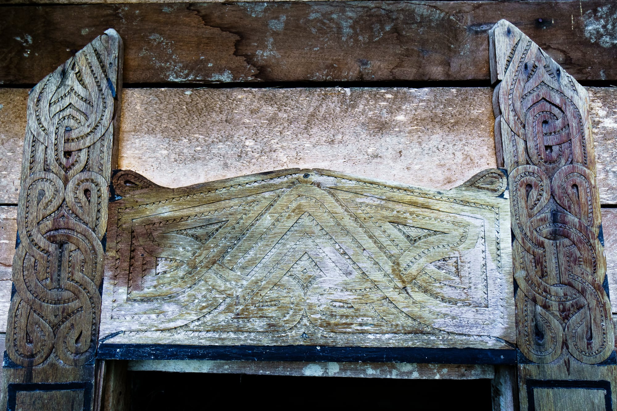 Weathered wooden boards carved with deeply cut interlacing bands and geometric patterns framing the top of a doorway