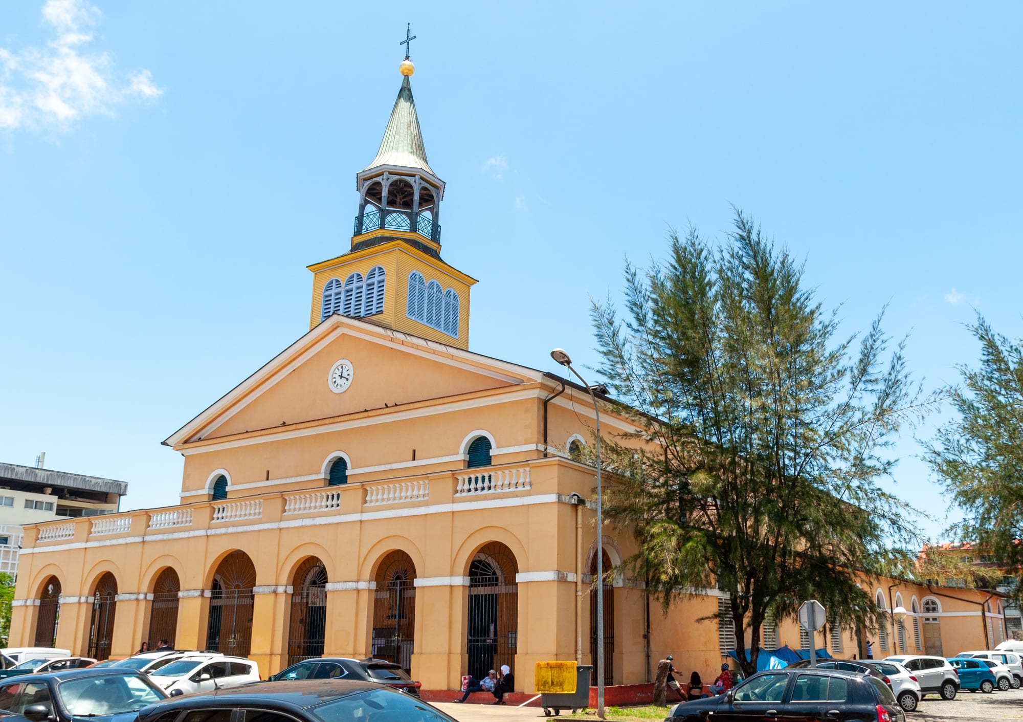 A pale orange church with white balustrades, arched doorways and a square bell tower topped by an open lantern and a cross, under a bright blue sky