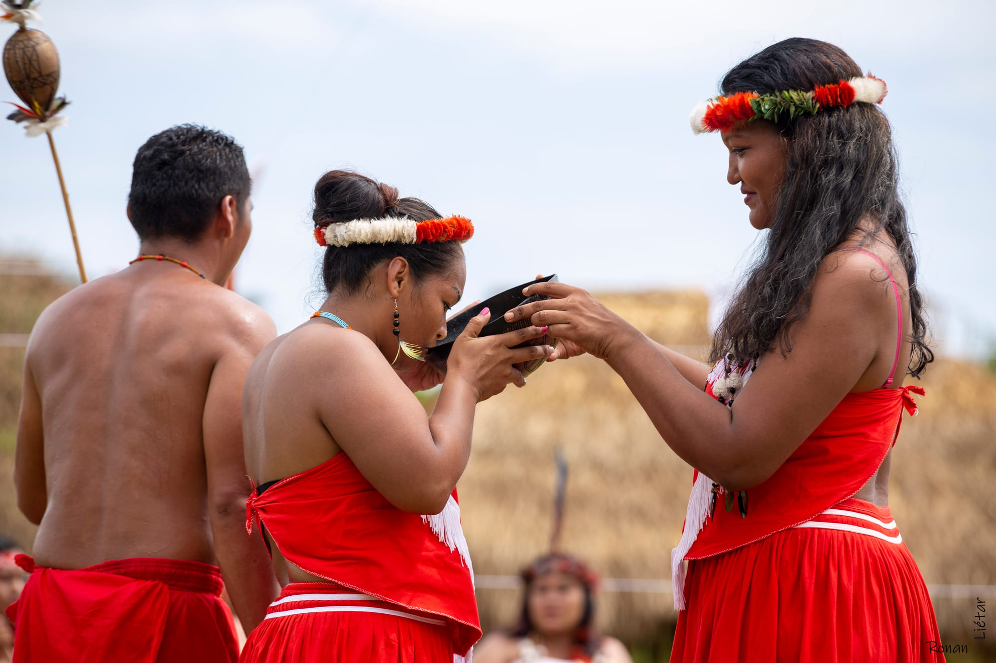 A woman in a red dress and a headdress of red and white feathers holds a dark bowl to the lips of another woman in red, who drinks from it, with a shirtless man in a red wrap beside them