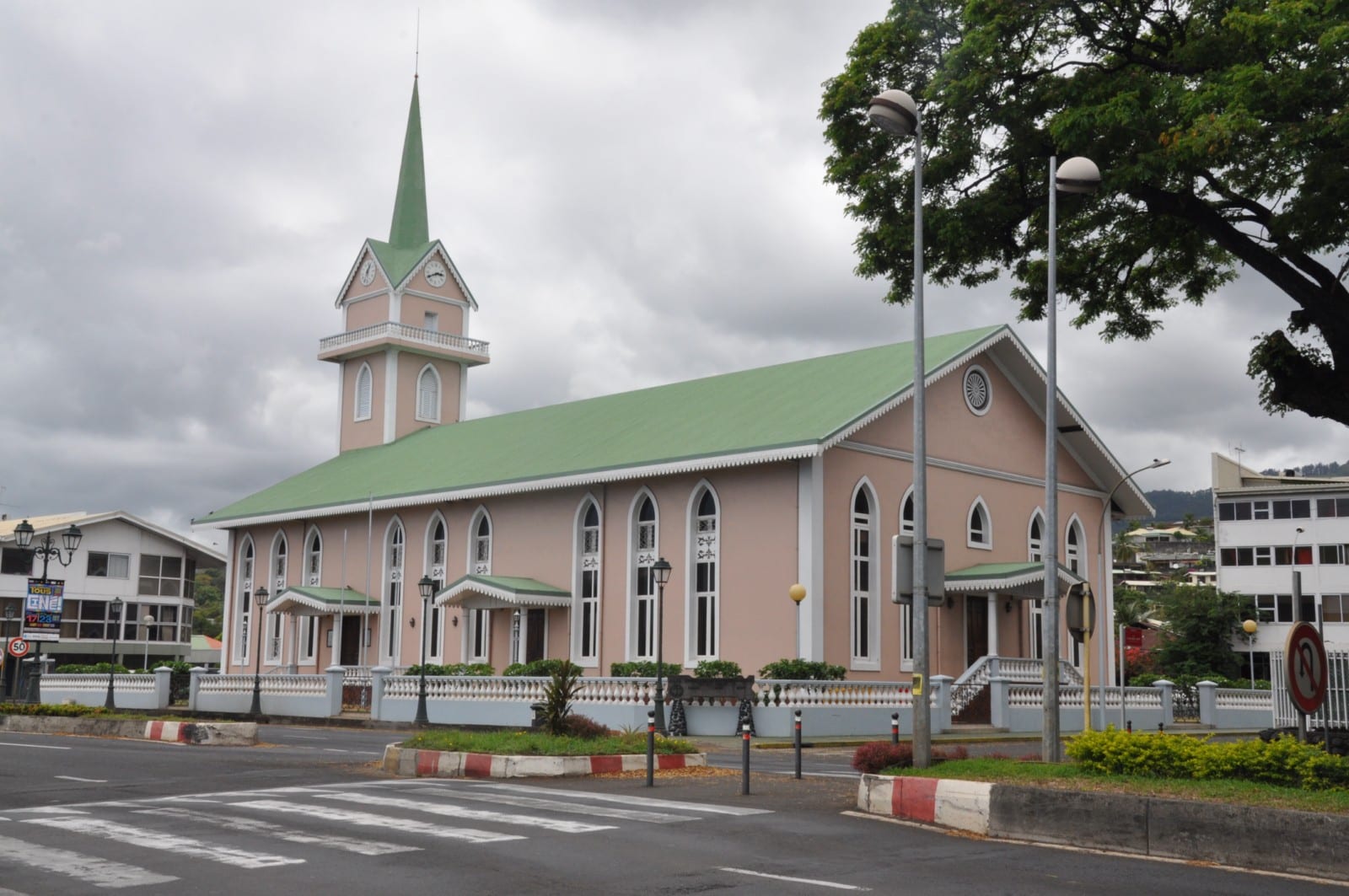 A large pink church with a green roof, tall arched white-framed windows and a clock tower with a green spire, behind a low white balustrade on a street corner