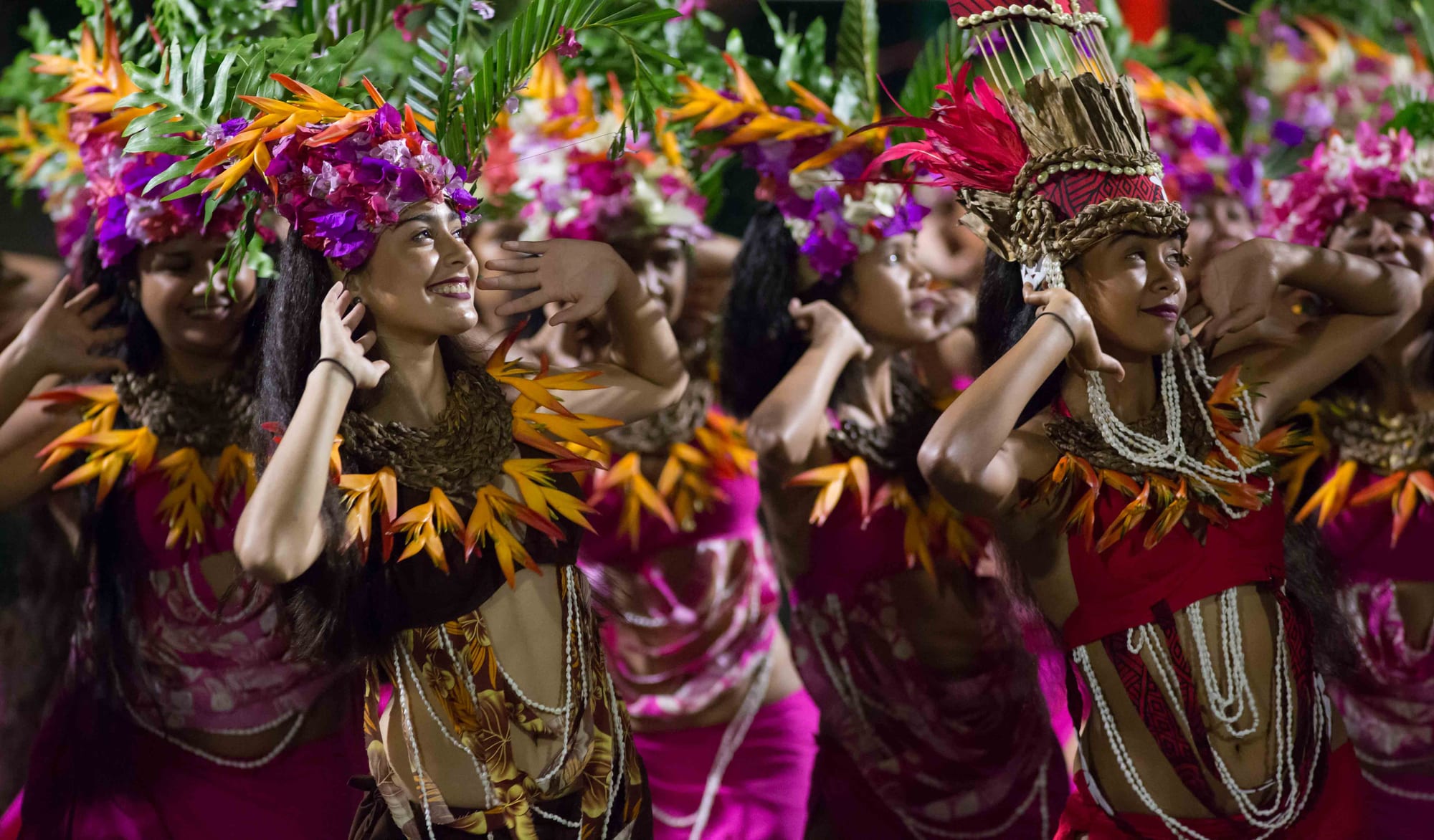 A line of women dancers in pink and patterned costumes with tall crowns of purple flowers, orange leaves and palm fronds, their hands raised beside their faces