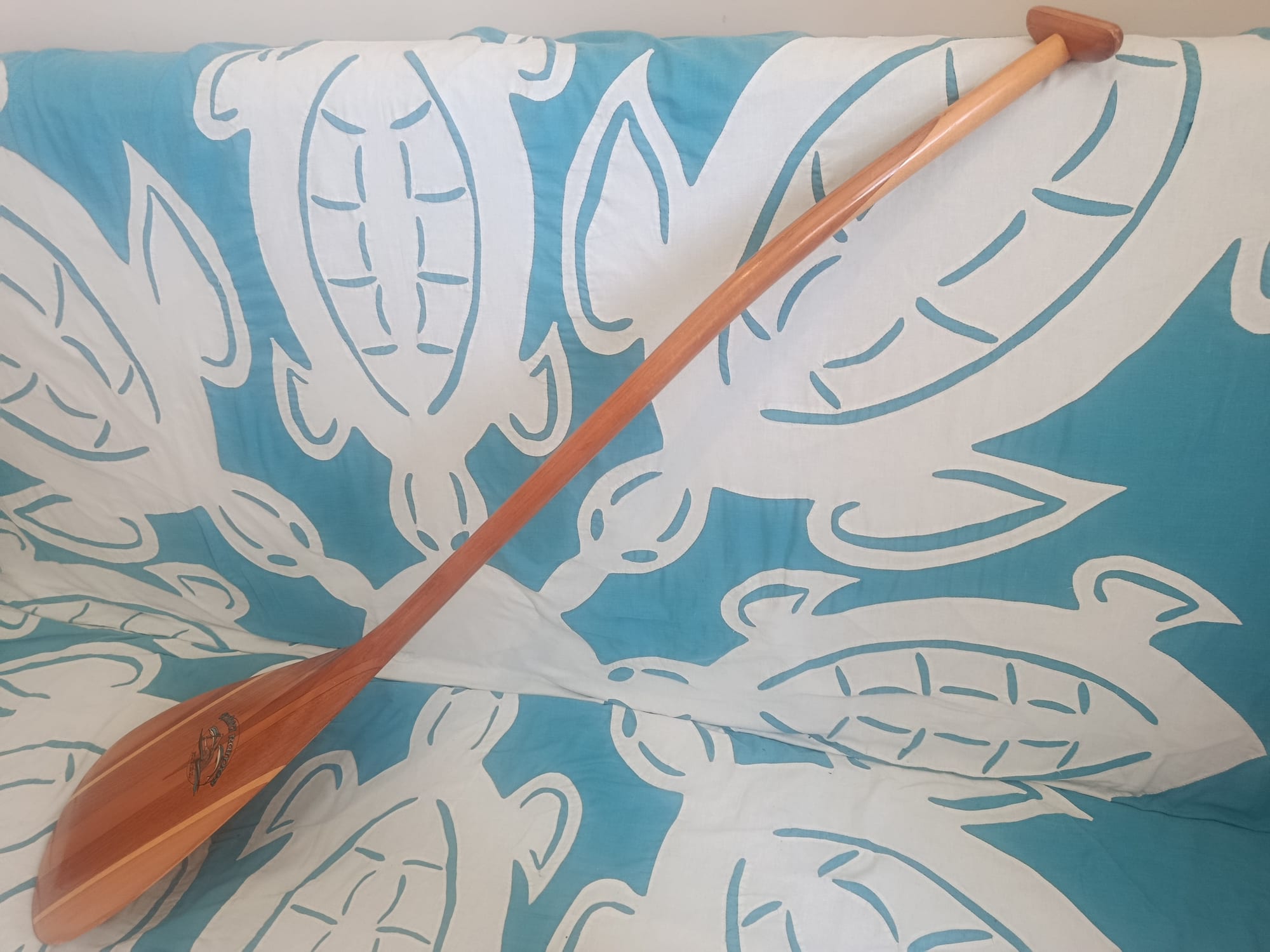 A varnished wooden canoe paddle laid diagonally across a turquoise and white appliqué quilt patterned with large stylised turtles