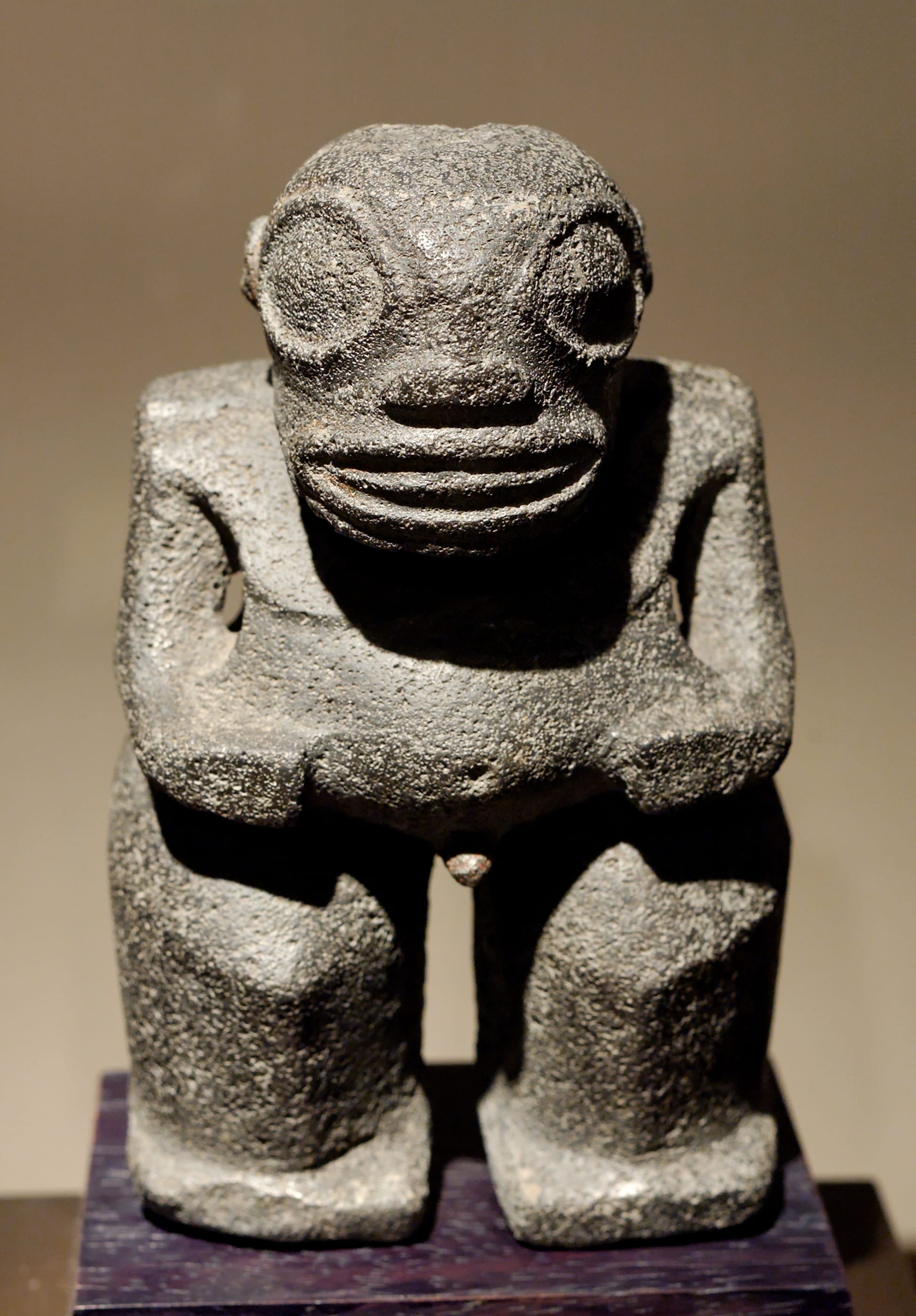 A small, squat grey stone figure with large round eyes, a wide banded mouth and hands resting on its belly, displayed on a dark plinth