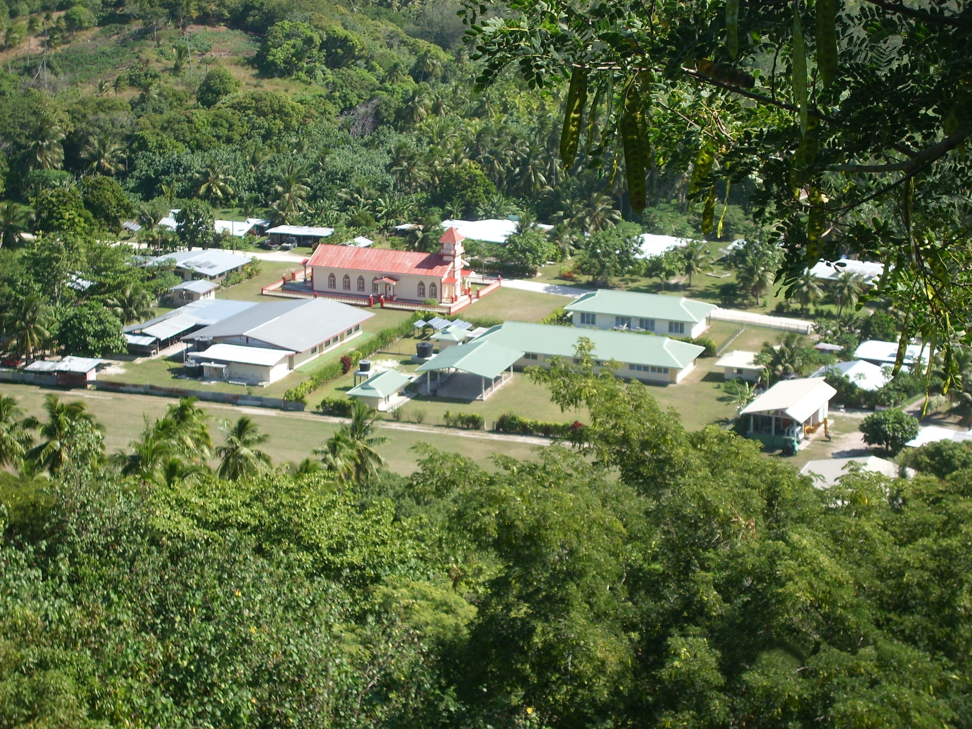 An aerial view of a small Polynesian village with a red-roofed church, houses with metal roofs, and palm trees on the island of Maiao