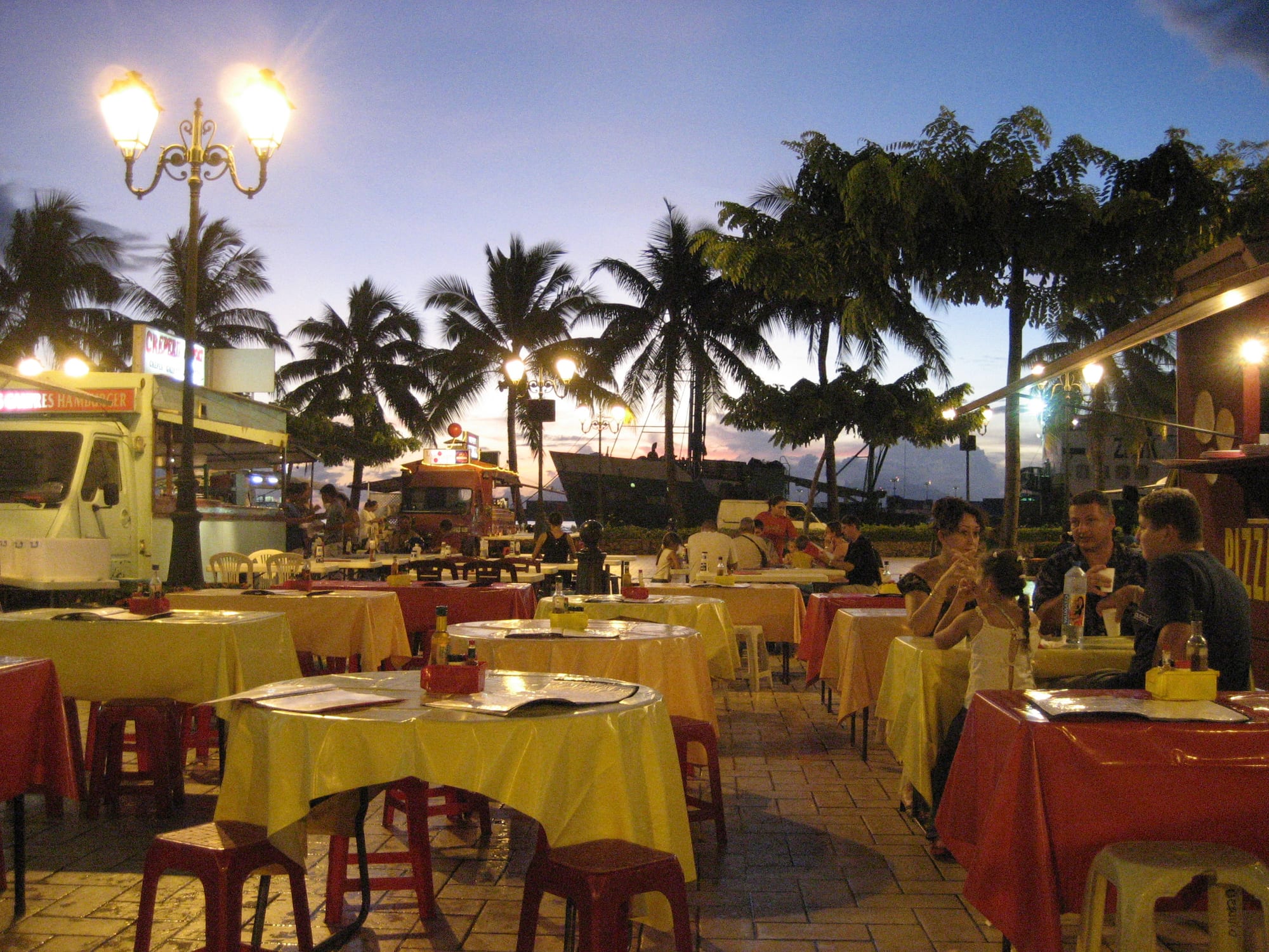Outdoor tables with red and yellow tablecloths set up beside food trucks on the Papeete waterfront at dusk, with palm trees and a harbour behind