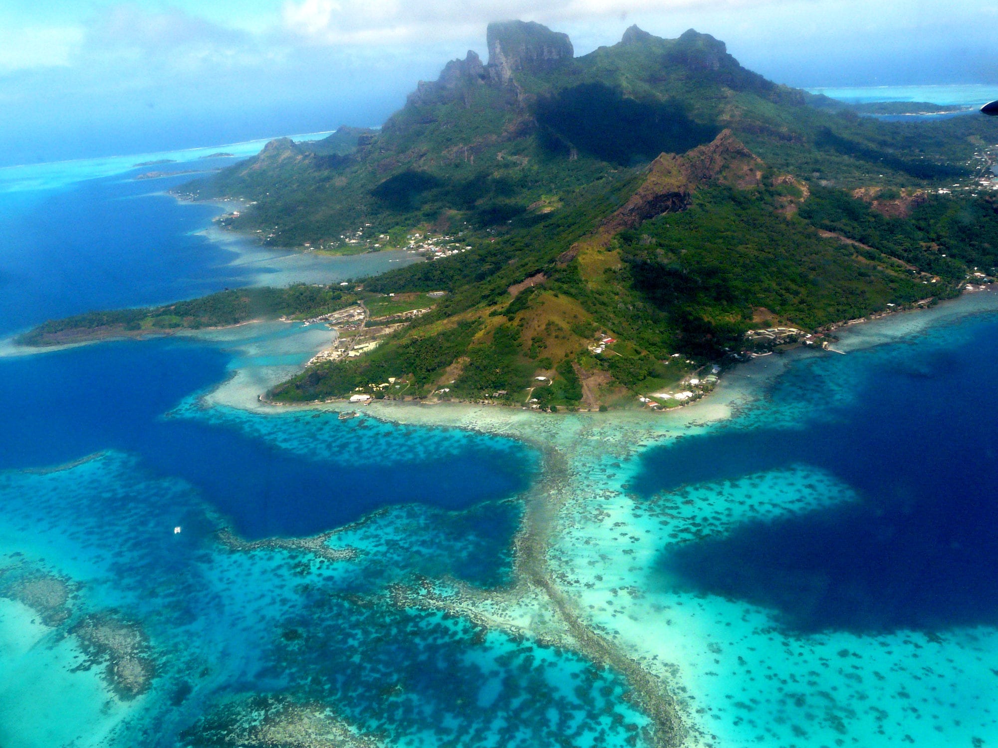A jagged green volcanic peak ringed by a turquoise lagoon and a reef, seen from the air