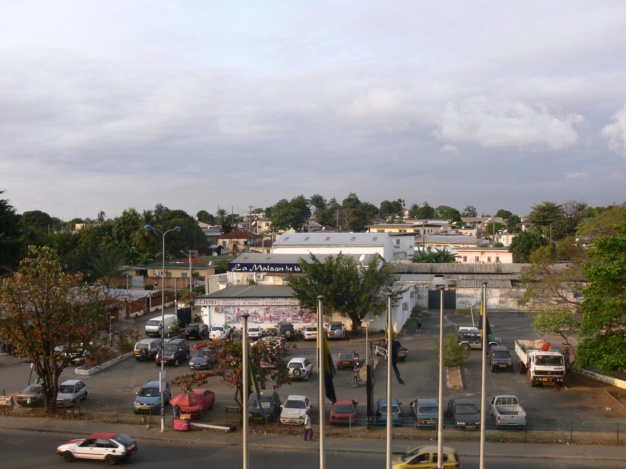 An elevated daytime view of a Libreville street with houses, shopfronts and parked cars under a partly cloudy sky