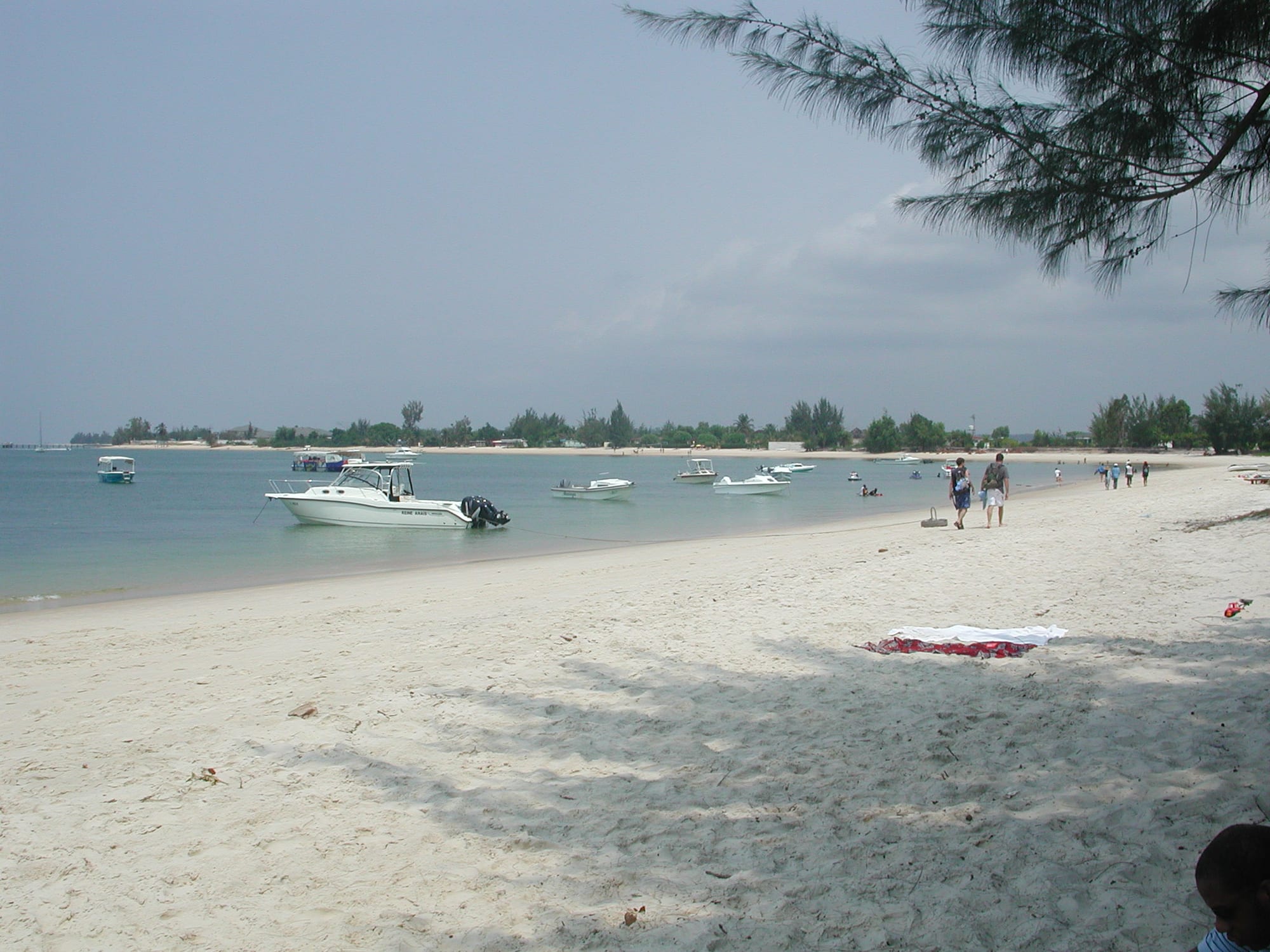 People walking along a white sand beach at Pointe Denis with small boats anchored just offshore