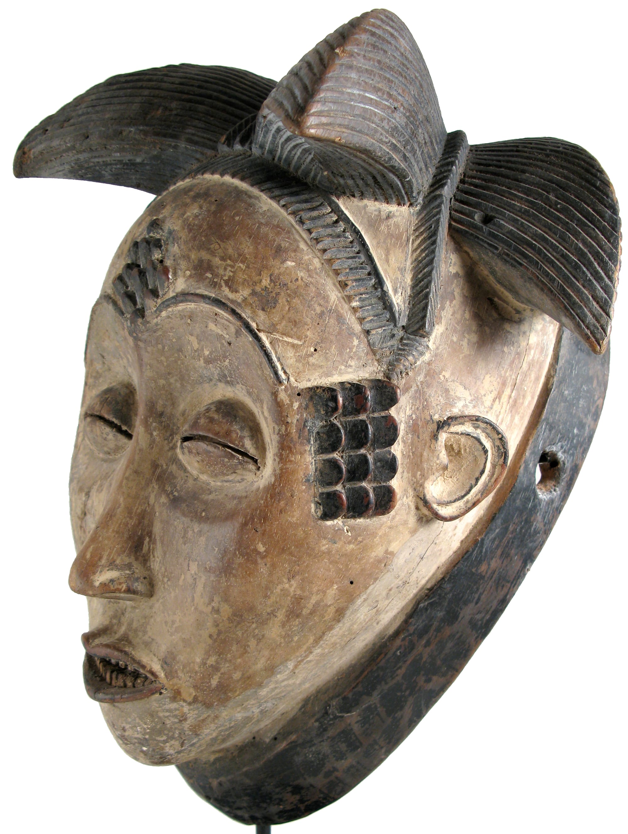 A carved wooden mask with a pale whitened face, narrow slit eyes, raised dark marks on the forehead and temple, and a tall dark crested hairstyle