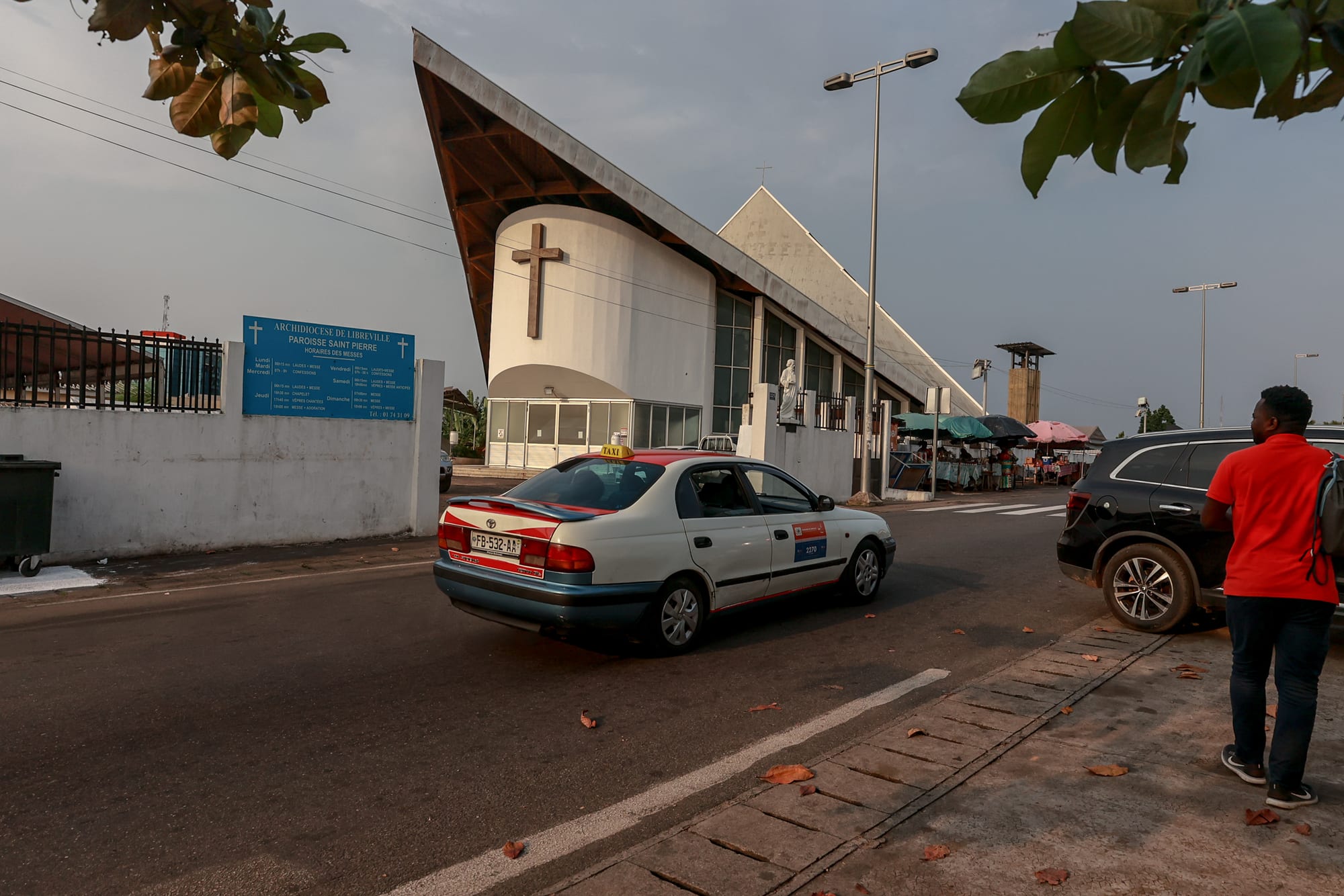 A modern white church with a steeply sloping roof and a large cross on its curved front wall, seen from a street where a taxi passes and a man walks along the pavement
