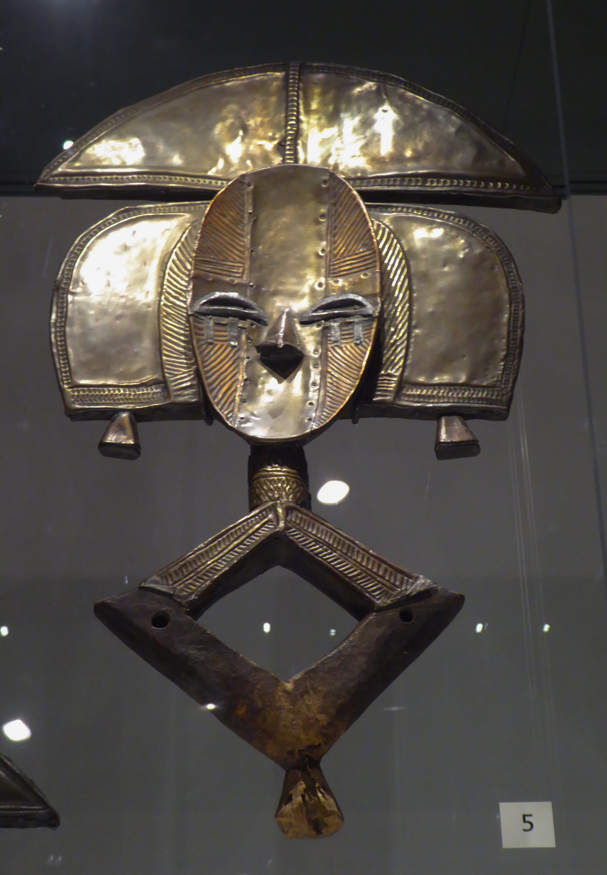 A flat, stylised figure with an oval face covered in polished brass and copper sheets, a wide crescent headdress and side panels, and a diamond-shaped wooden base, displayed in a glass case