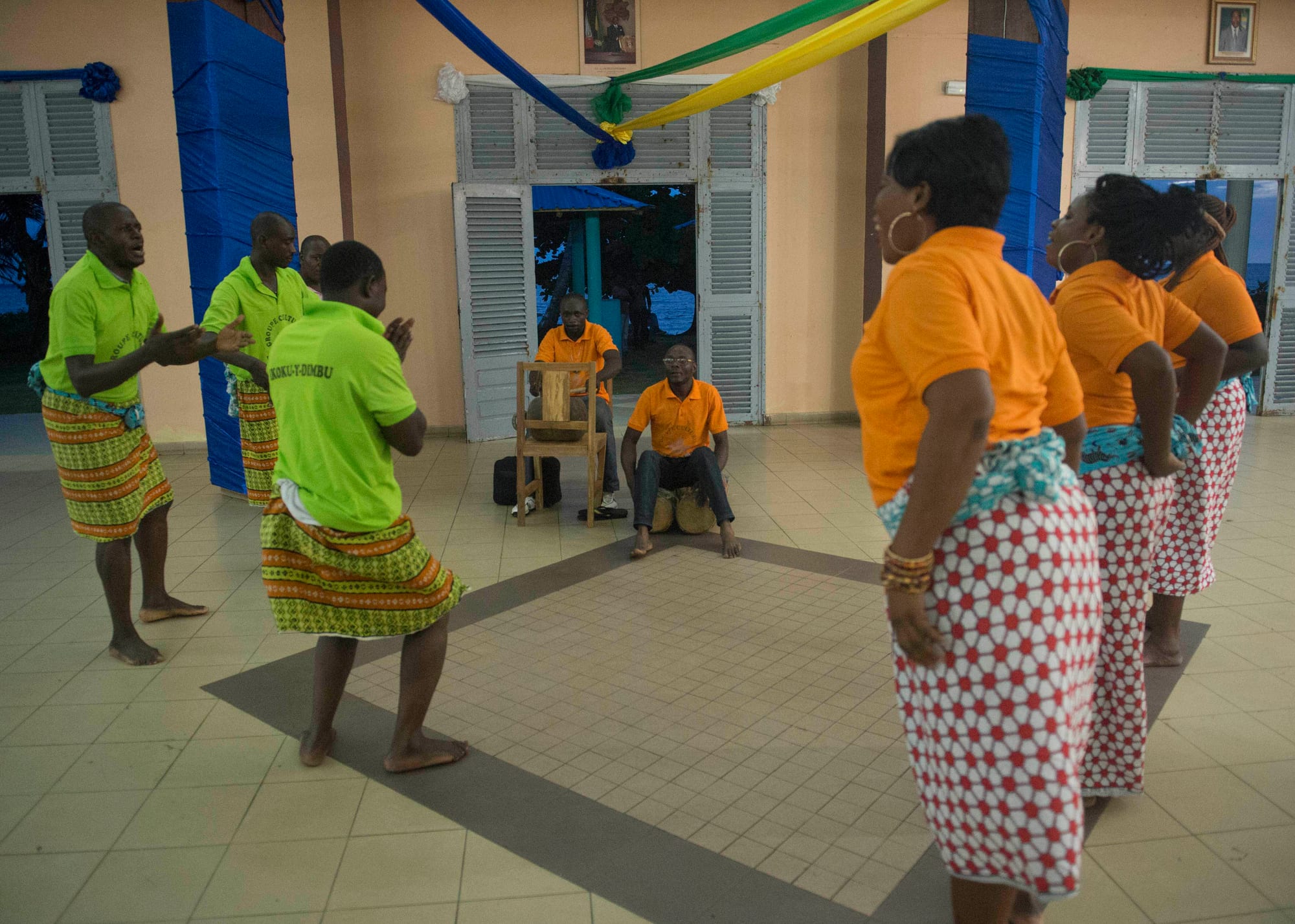 Men in green shirts and patterned wrap skirts and women in orange shirts and red and white wrap skirts dance barefoot on a tiled floor, while a man seated behind them plays a drum