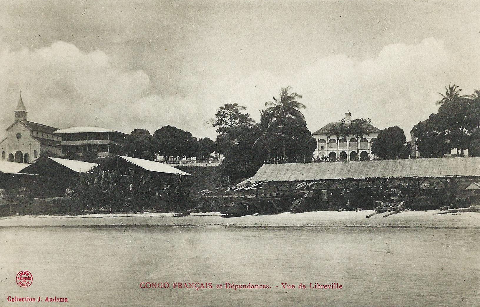 A sepia colonial-era postcard of a tropical waterfront seen from the water, with a church, long open-sided sheds on the beach, palms and a large arcaded building on the rise behind