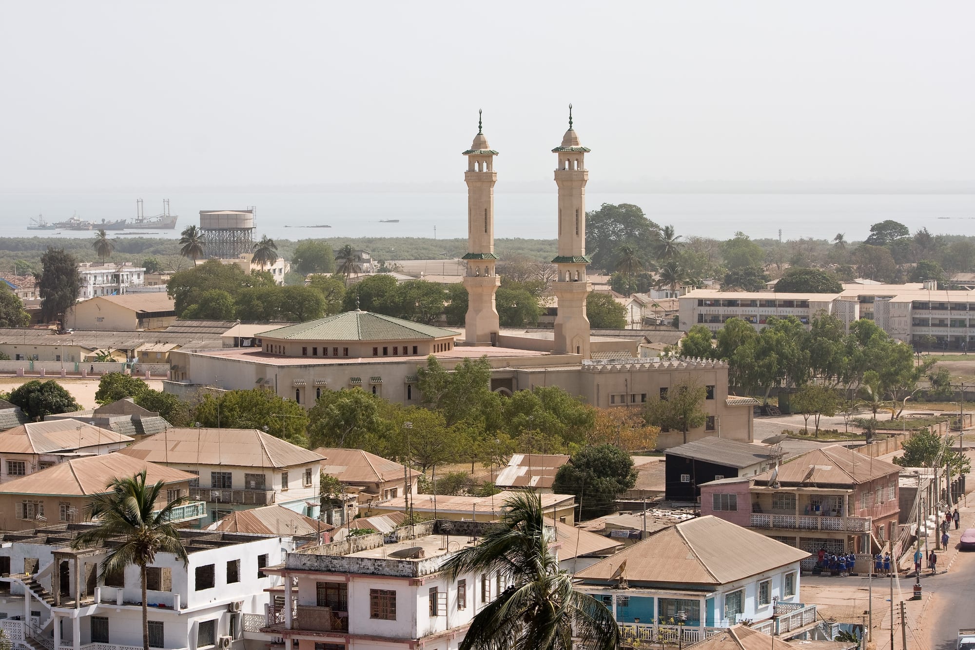 A large mosque with two tall pale minarets and a green-tiled roof rising above low buildings, palm trees and tin roofs, with the river estuary and a ship on the horizon