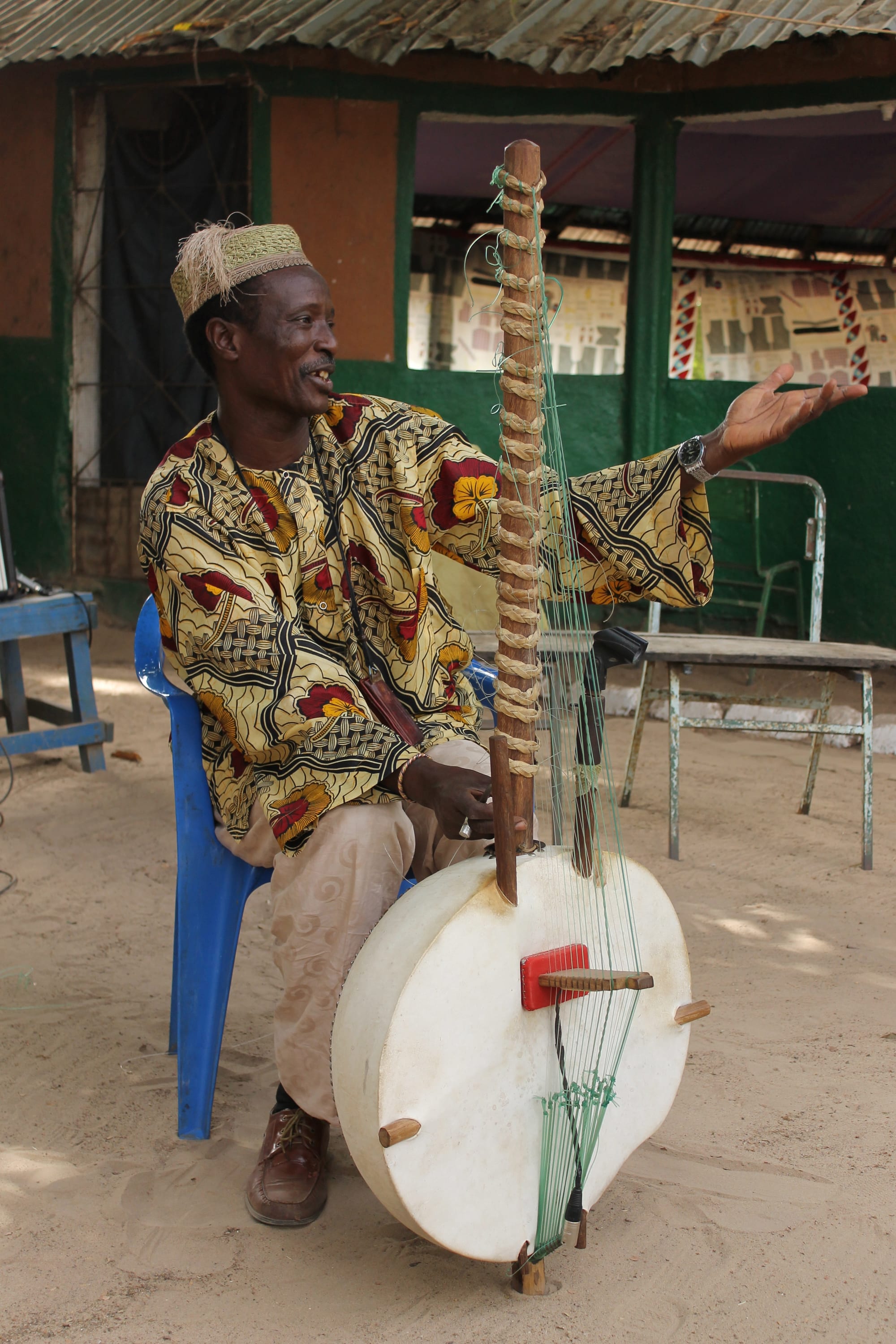 A man in a patterned yellow and red gown and an embroidered cap sits on a blue plastic chair holding a kora, a tall-necked harp with a large round cowhide-covered body, and gestures with his free hand