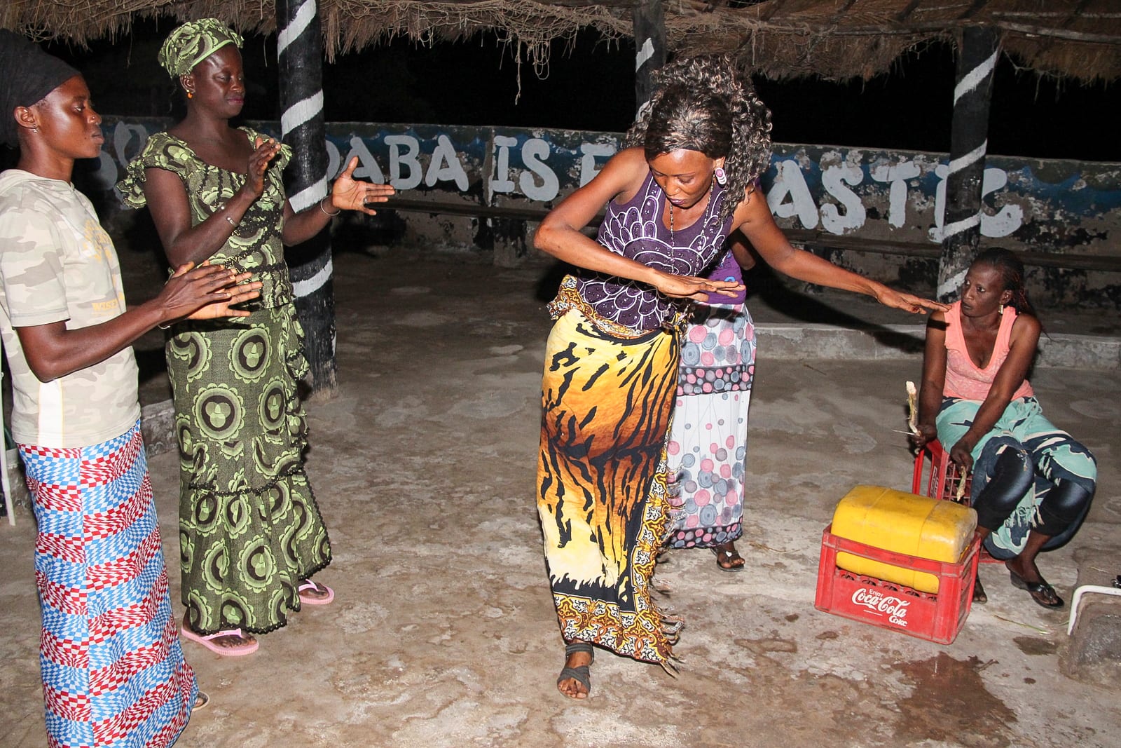 A woman in a patterned wrap skirt dances with her arms outstretched on a concrete floor under a thatched shelter, while other women in colourful dresses clap and one sits on a plastic crate