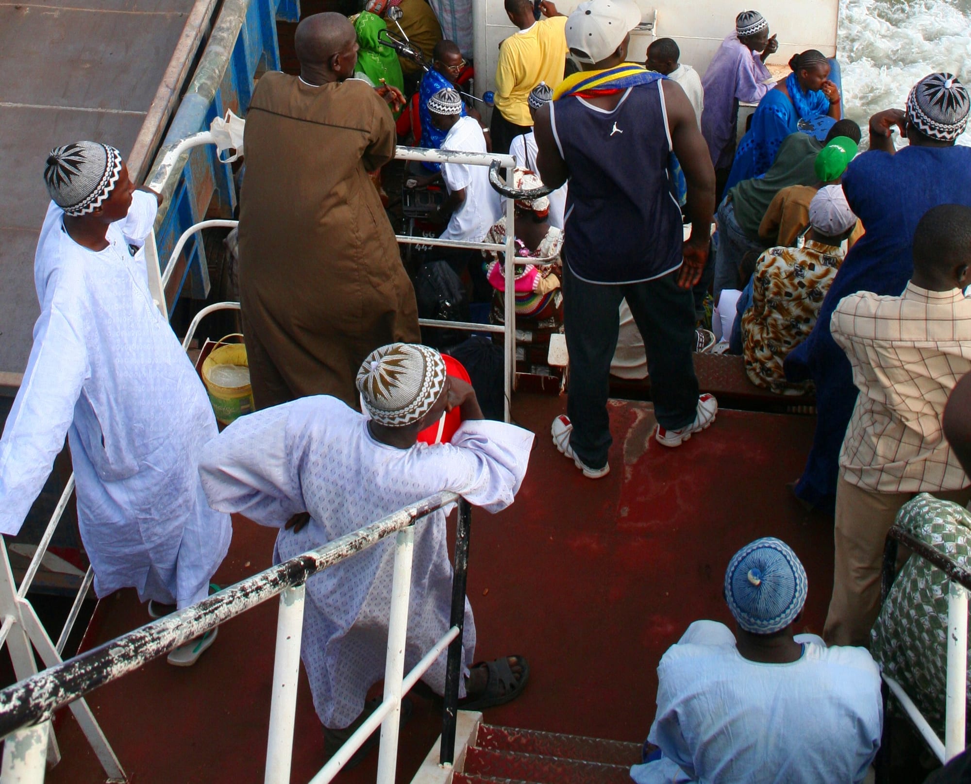 A crowd of passengers in colourful dress standing on the deck of the Banjul-Barra ferry crossing the river