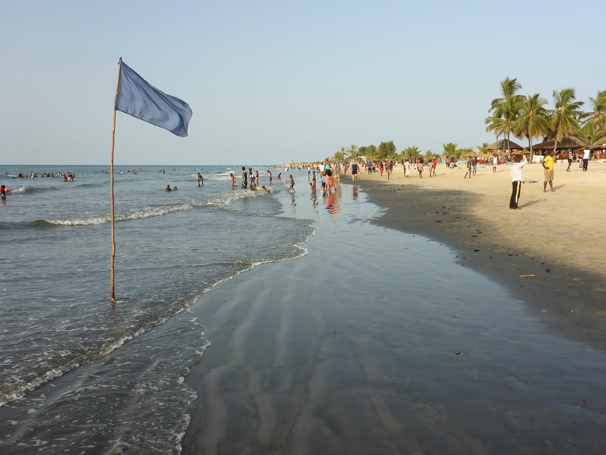 A wide sandy beach at Kololi with a safety flag, thatched umbrellas and palm trees, and people walking along the shore