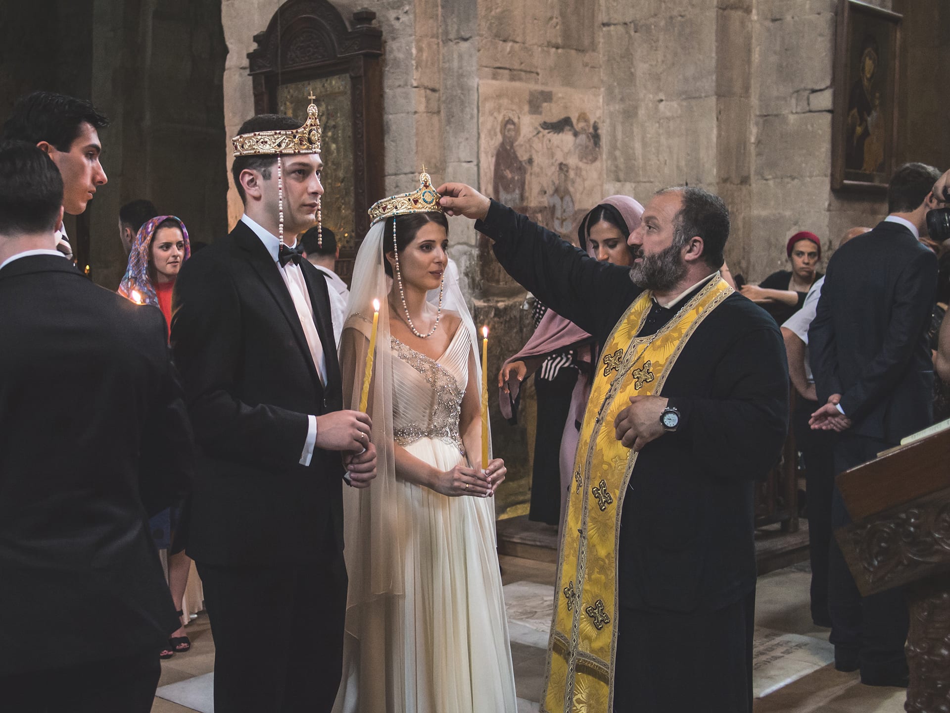 A bride and groom wearing jewelled crowns and holding lit candles stand in a stone church while a bearded priest in a gold stole rests his hand on the bride's crown, with guests behind them